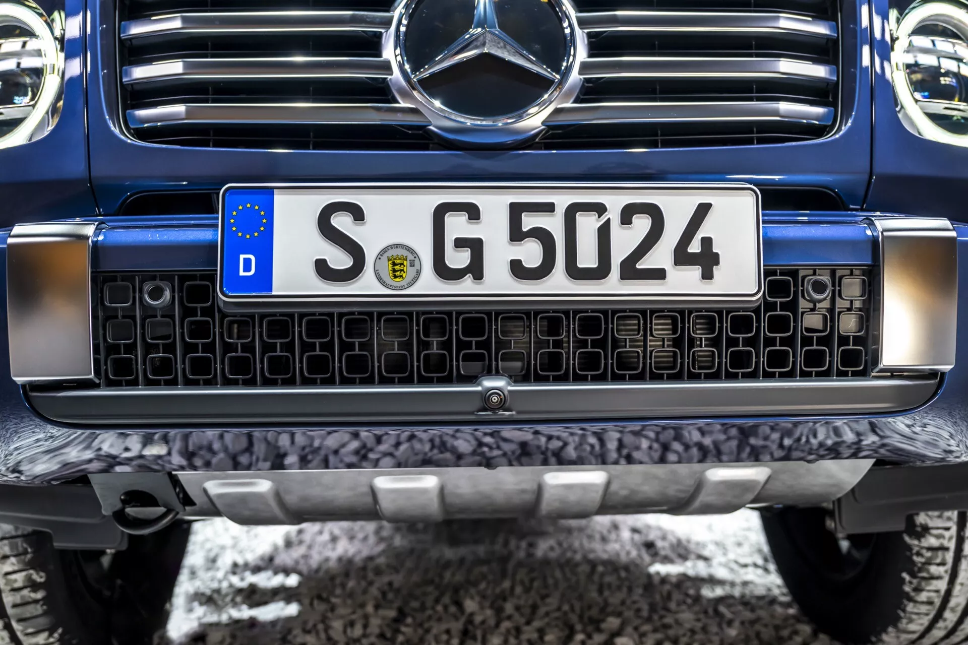 Zbliżony widok na przednią część Mercedes-Benz klasy G 2024, przedstawiający grill z czarnymi prostokątami z chromowanymi obramowaniami, europejską tablicę rejestracyjną, solidny przedni zderzak z metalową płytą osłonową i centralny emblemat Mercedes-Benz z potencjalną technologią sensorów, pomalowany na ciemnoniebiesko, wskazujący na luksusowy lub wysokowydajny model SUV w idealnym stanie.
