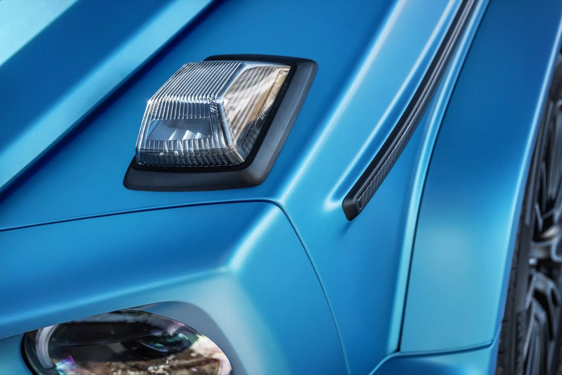 Szczegółowy zbliżenie Mercedes-Benz G-Klasse 2024 z wlotem powietrza na masce, kanciastymi liniami nadwozia, metalicznym niebieskim lakierem oraz bocznym wlotem powietrza, pokazujące elementy designu i technologii