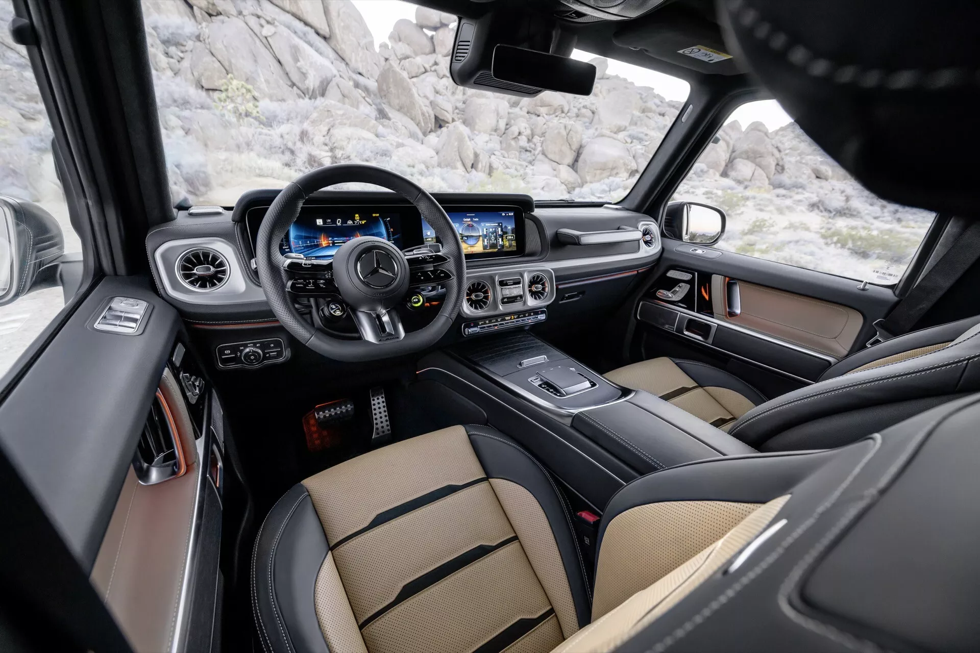 Widok wnętrza Mercedes-Benz G-Klasse 2024 (465) ukazujący kokpit kierowcy z wielofunkcyjną kierownicą, cyfrowym zegarem, dużym systemem informacyjno-rozrywkowym z ekranem dotykowym, luksusowymi siedzeniami z dwukolorowej skóry i zaawansowanymi funkcjami bezpieczeństwa z perspektywy pasażera tylnej