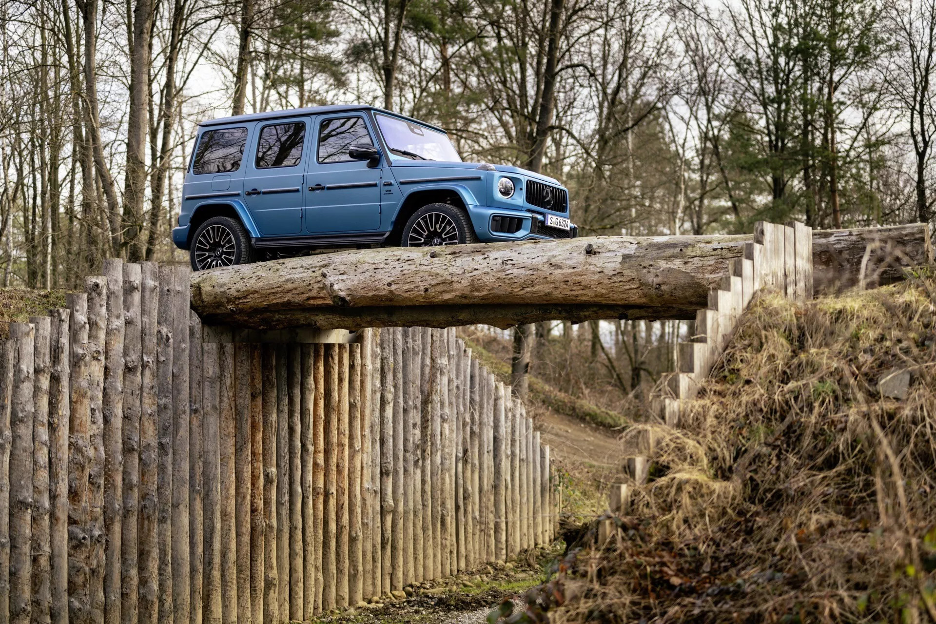 Niebieskie SUV Mercedes-Benz Klasy G z 2024 roku na drewnianym moście podkreślające zdolności terenowe, profil boczny z dużym prześwitem, w lesistym otoczeniu