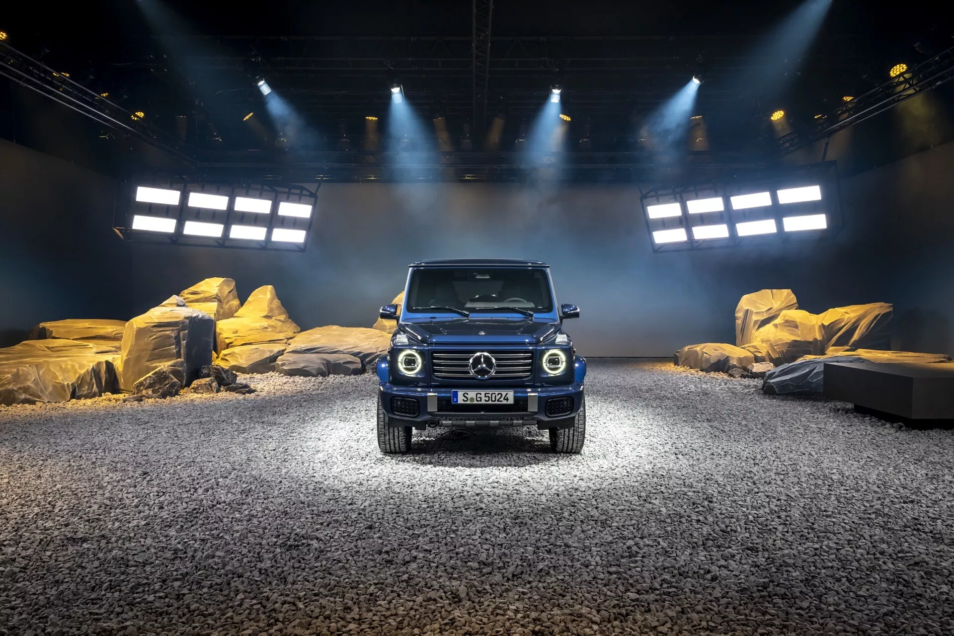 SUV Mercedes-Benz Klasy G 2024 na symulowanej scenie zewnętrznej z oświetleniem punktowym, pokazujący przedni grill, okrągłe reflektory i solidną konstrukcję odpowiednią do warunków terenowych