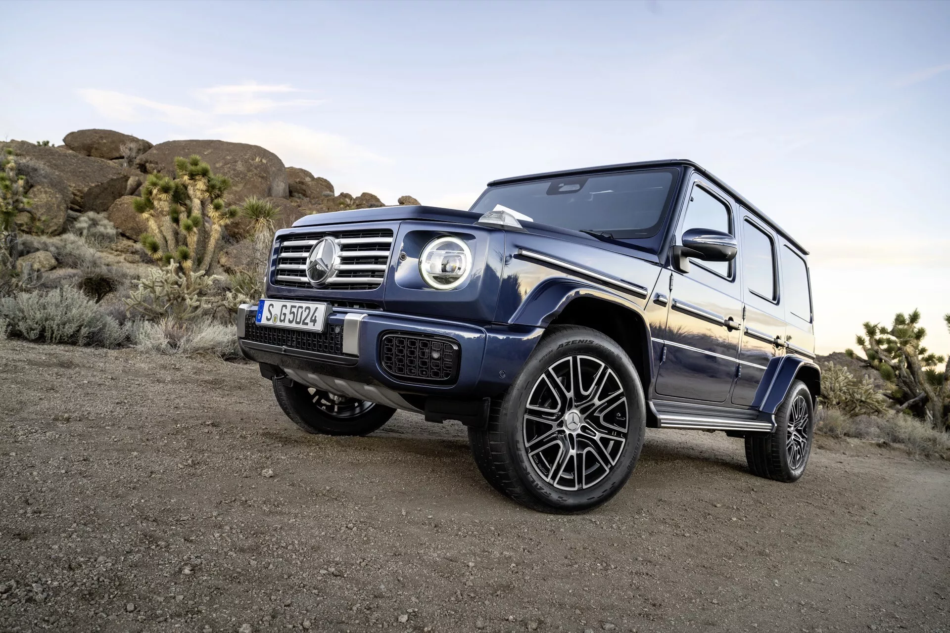 Dynamiczny kątowy zdjęcie SUV-a Mercedes-Benz Klasy G 2024 ukazujące jego potężną postawę i szczegółowe elementy projektu, takie jak okrągłe reflektory, przednia kratka z emblematem i muskularne nadkola, na tle terenowym w miękkim wieczornym świetle