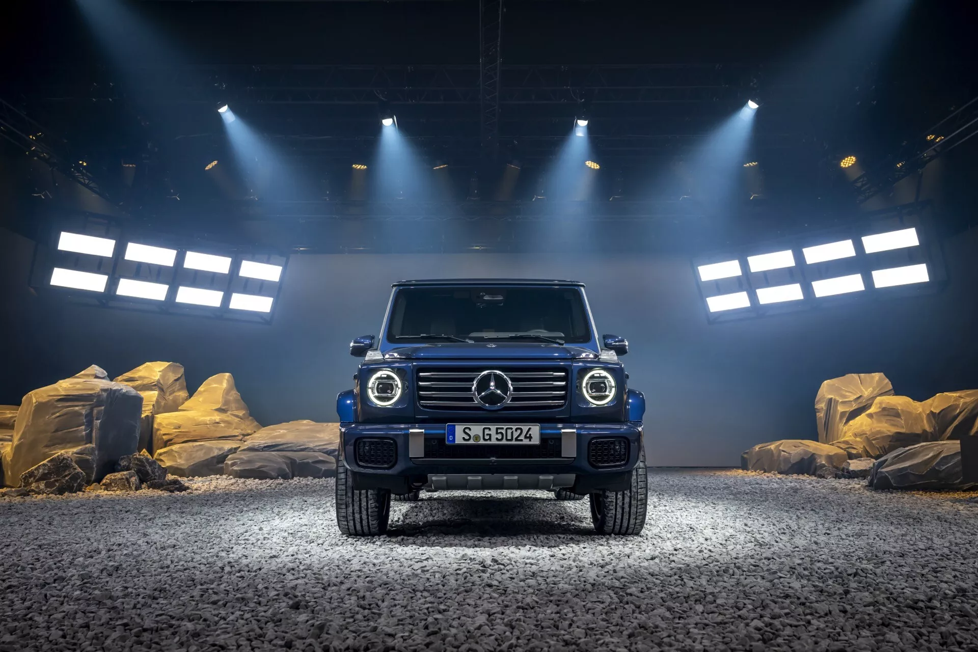 Luksusowy SUV terenowy Mercedes-Benz Klasa G (G-Klasse) 2024 z dramatycznym oświetleniem, podkreślającym wysoki prześwit, ikoniczną grillę z emblematem, okrągłe reflektory LED, prostokątne światła do jazdy dziennej, wytrzymały przedni zderzak i muskularny design maski na żwirowej nawierzchni z przygodowym tłem studia