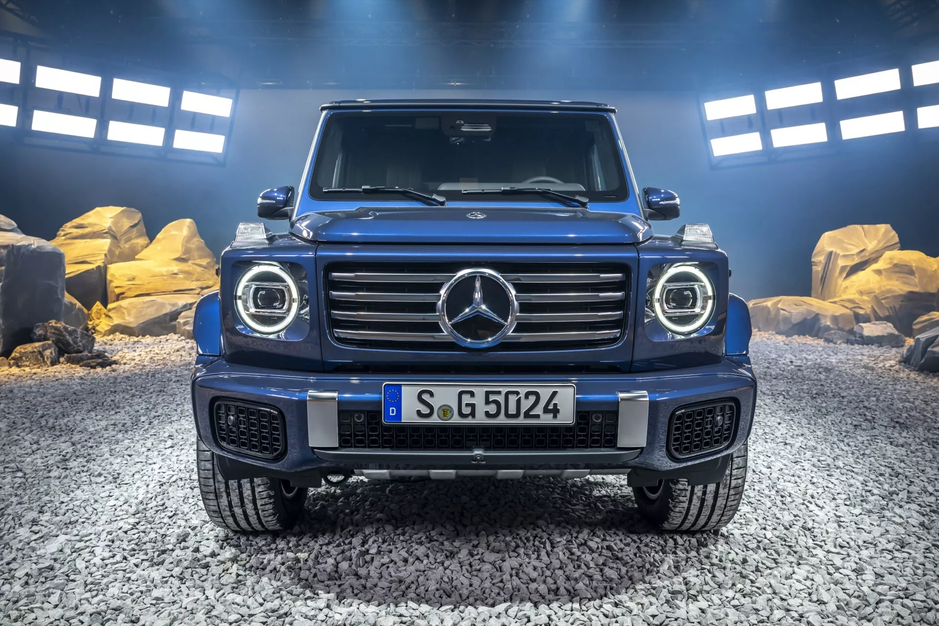 Przedni widok Mercedes-Benz Klasy G 2024 z ikoniczną kratą, reflektorami LED, robustnym designem maski oraz przednim zderzakiem gotowym do jazdy w terenie z płytą ochronną, uchwycony w naturalnym otoczeniu na zewnątrz