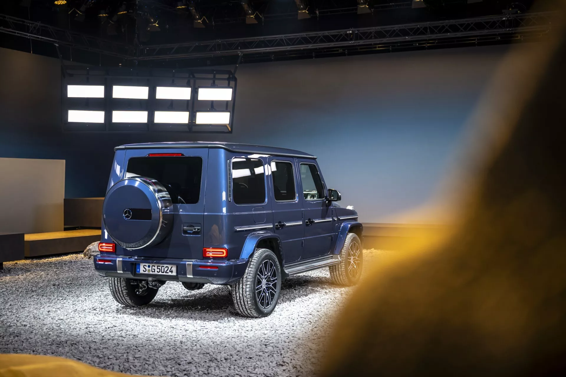 Boczny i częściowo tylny widok luksusowego SUV Mercedes-Benz Klasy G 2024, podkreślający cechy terenowego designu, aluminiowe felgi i przyciemniane szyby w scenerii wewnętrznej wystawy