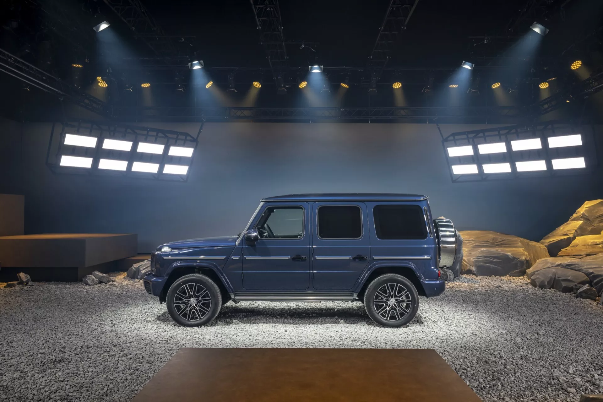 Boczny profil luksusowego SUV-a Mercedes-Benz G-Klasse 465 z 2024 roku na symulowanej wewnętrznej scenie terenowej, podkreślający estetykę designu, stabilność i zaawansowane funkcje technologiczne pod jasnym oświetleniem reflektorów