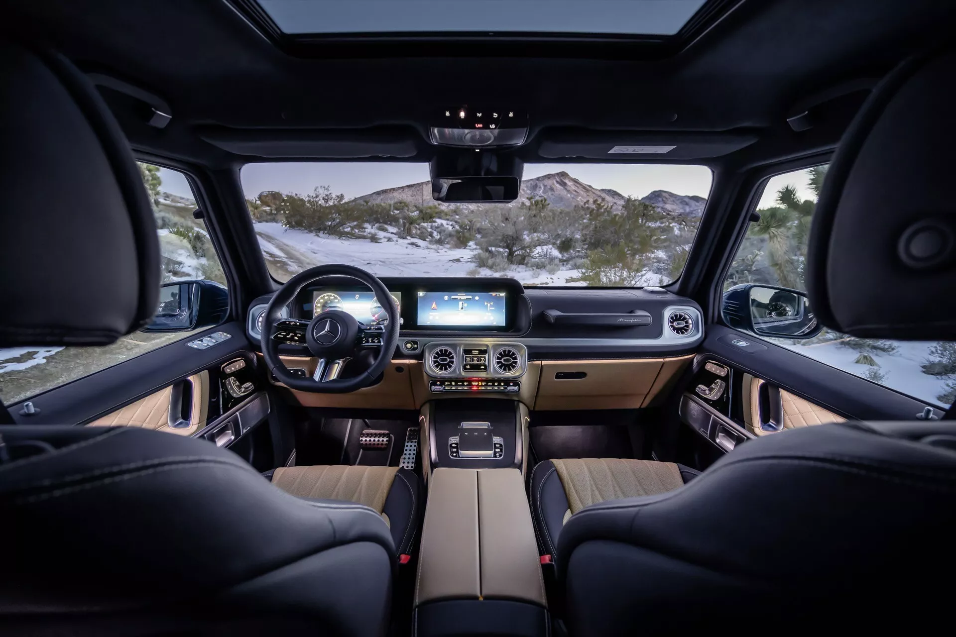 Widok wnętrza Mercedes-Benz Klasy G 2024 z perspektywy tylnego centrum, prezentujący wielofunkcyjną kierownicę z obszytą skórą, podwójne wyświetlacze cyfrowe na desce rozdzielczej, konsolę środkową z panelem dotykowym, dźwignię zmiany biegów automatycznych, czarne skórzane fotele z elektroniczną regulacją, akcenty drewniane, oświetlenie ambientowe i wyraźny widok przez przednią szybę