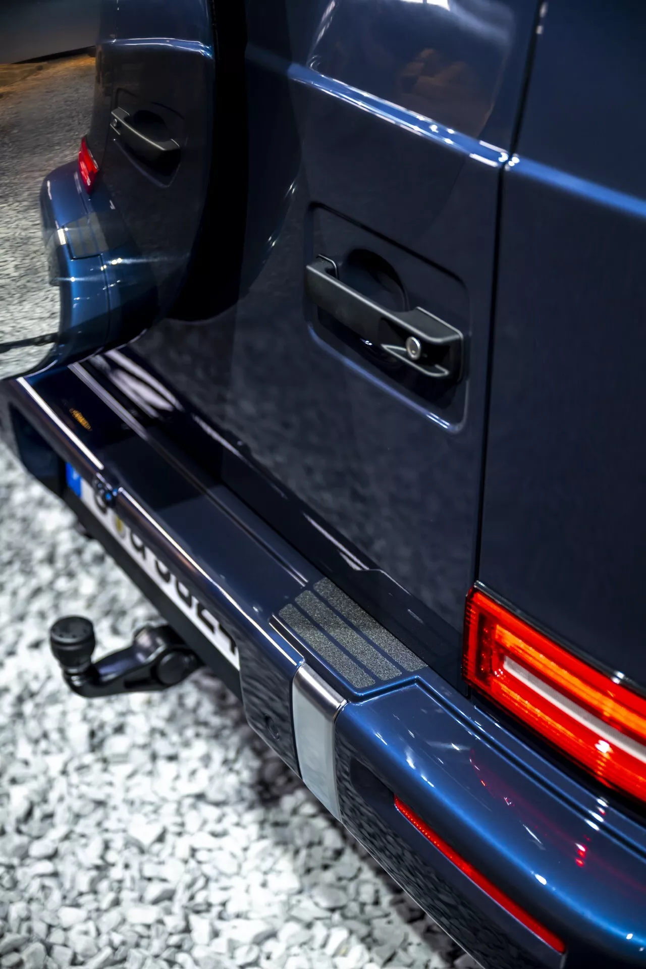 Zbliżony widok kątowy podkreślający tylną część Mercedes-Benz G-Klasse 2024, prezentujący prawą stronę tylnej lampy, zderzaka i detal drzwi z oświetleniem LED i funkcjami holowania.