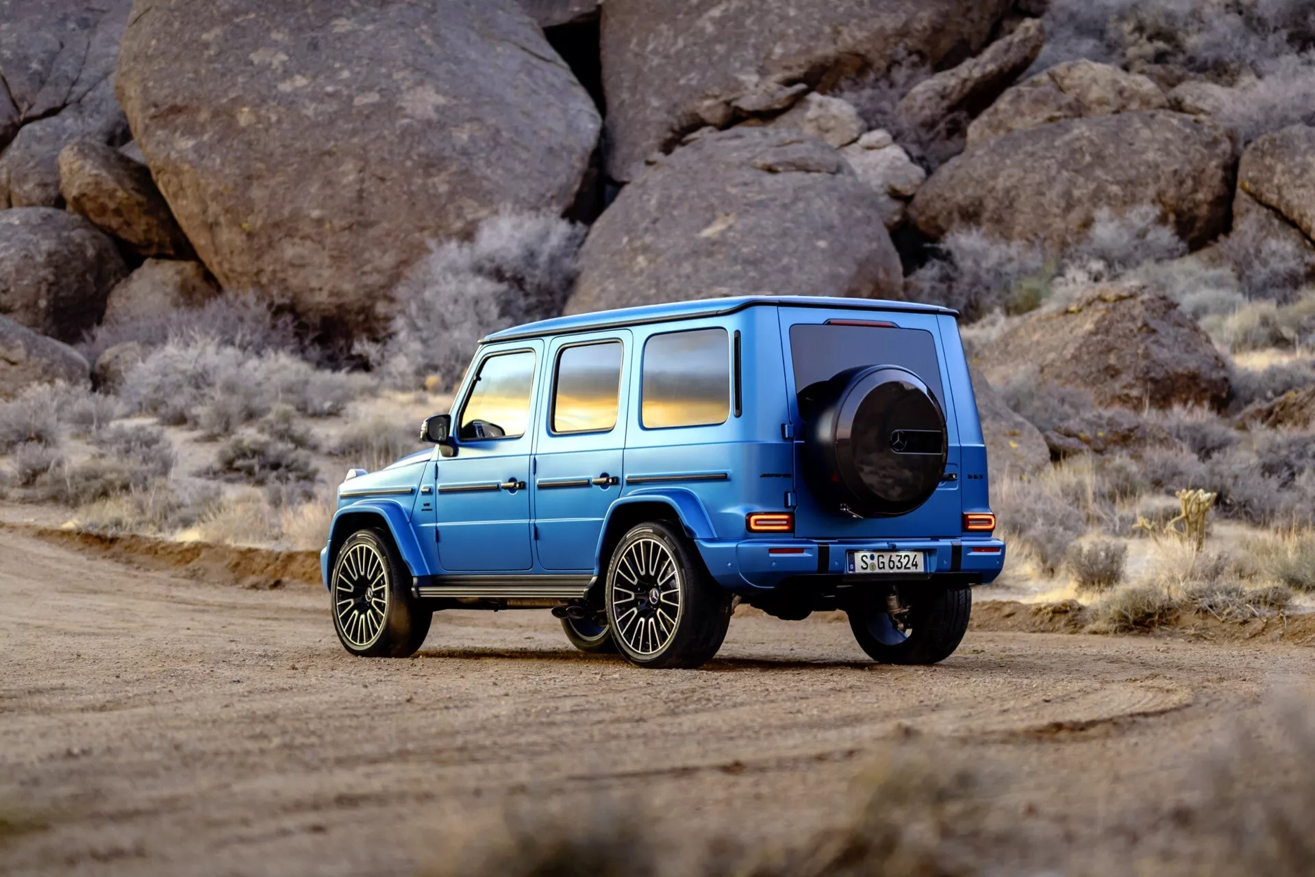 Luksusowe niebieskie SUV Mercedes-Benz Klasy G (G-Klasse) 465 zaparkowane na drodze żwirowej z tłem dużych, szorstkich skał, podkreślające kąt boczny tyłu, koło zapasowe i felgi aluminiowe w otoczeniu przypominającym pustynię.