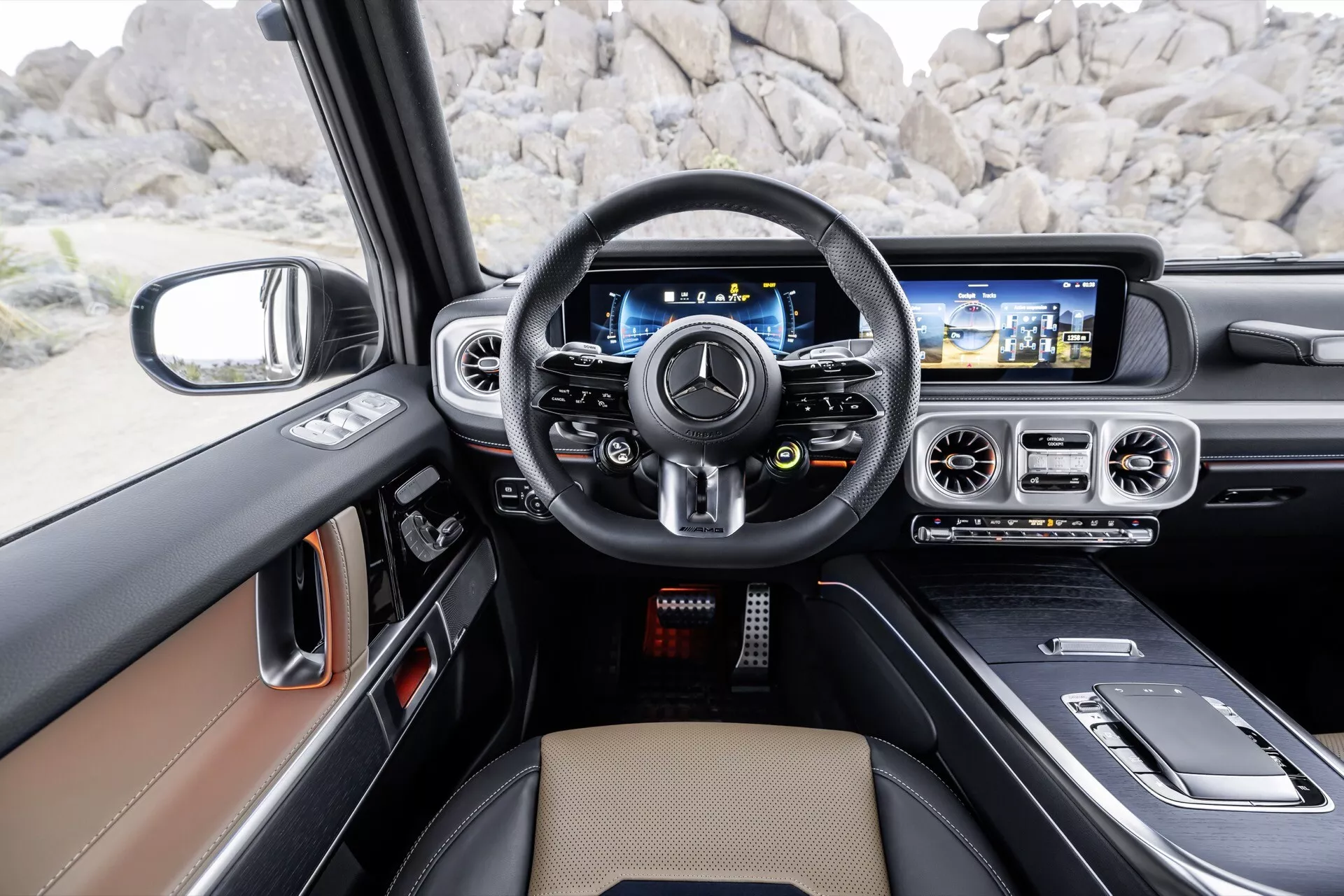 Widok wnętrza Mercedes-Benz klasy G (G-Klasse) 2024 z perspektywy fotela kierowcy pokazujący wielofunkcyjną kierownicę pokrytą skórą, cyfrowy zestaw wskaźników, szeroki ekran systemu infotainment z nawigacją oraz luksusowy wnętrze w kolorach jasnobrązowym i czarnym z akcentami z metalu szczotkowanego i możliwymi elementami z drewna