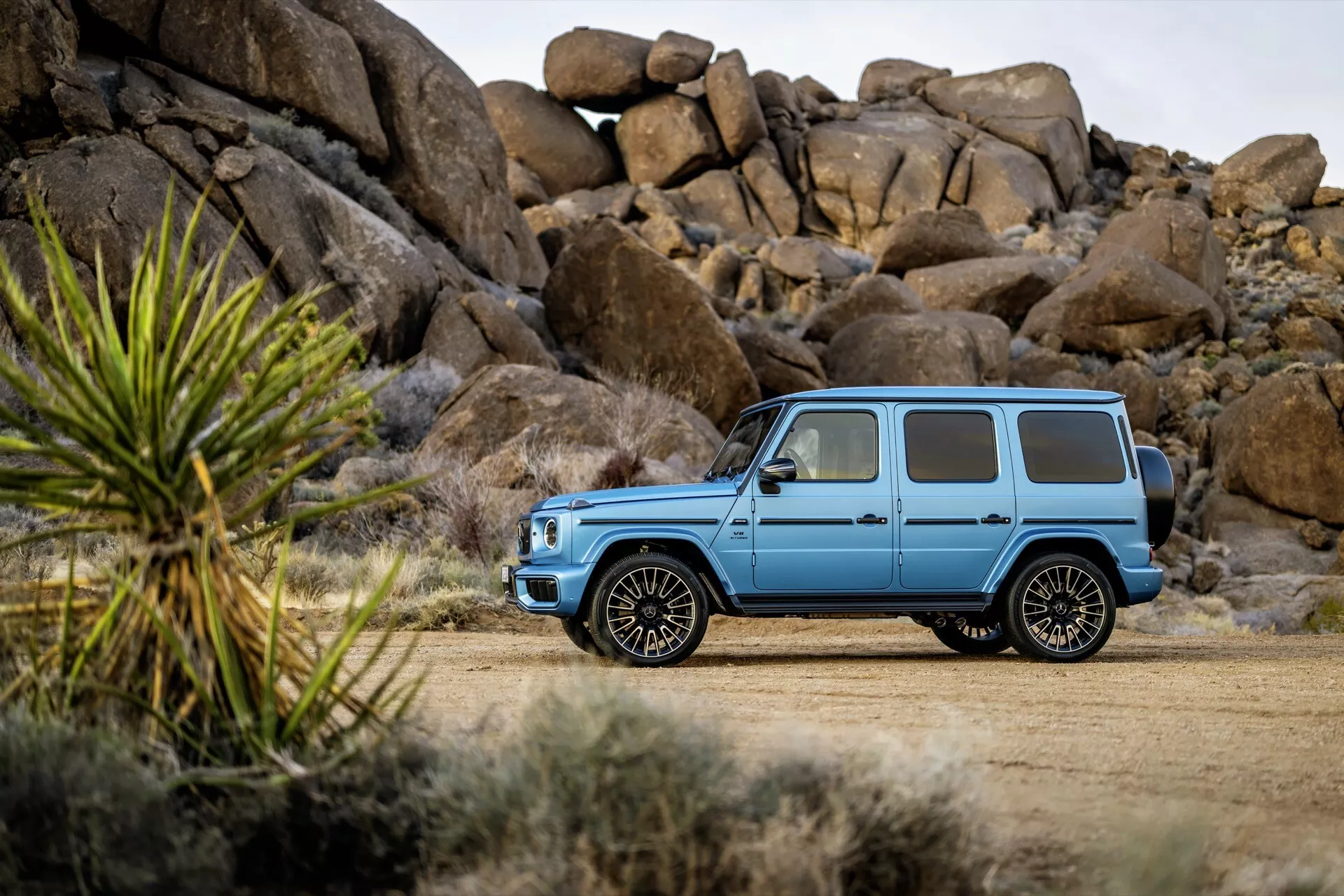 Luksusowy SUV Mercedes-Benz Klasa G (G 465) z 2024 r. w jasnoniebieskim matowym kolorze, prezentujący swój solidny design i wysokie prześwit na drodze gruntowej, z zaawansowanymi kołami ze stopu i przyciemnianymi szybami na tle surowego krajobrazu.
