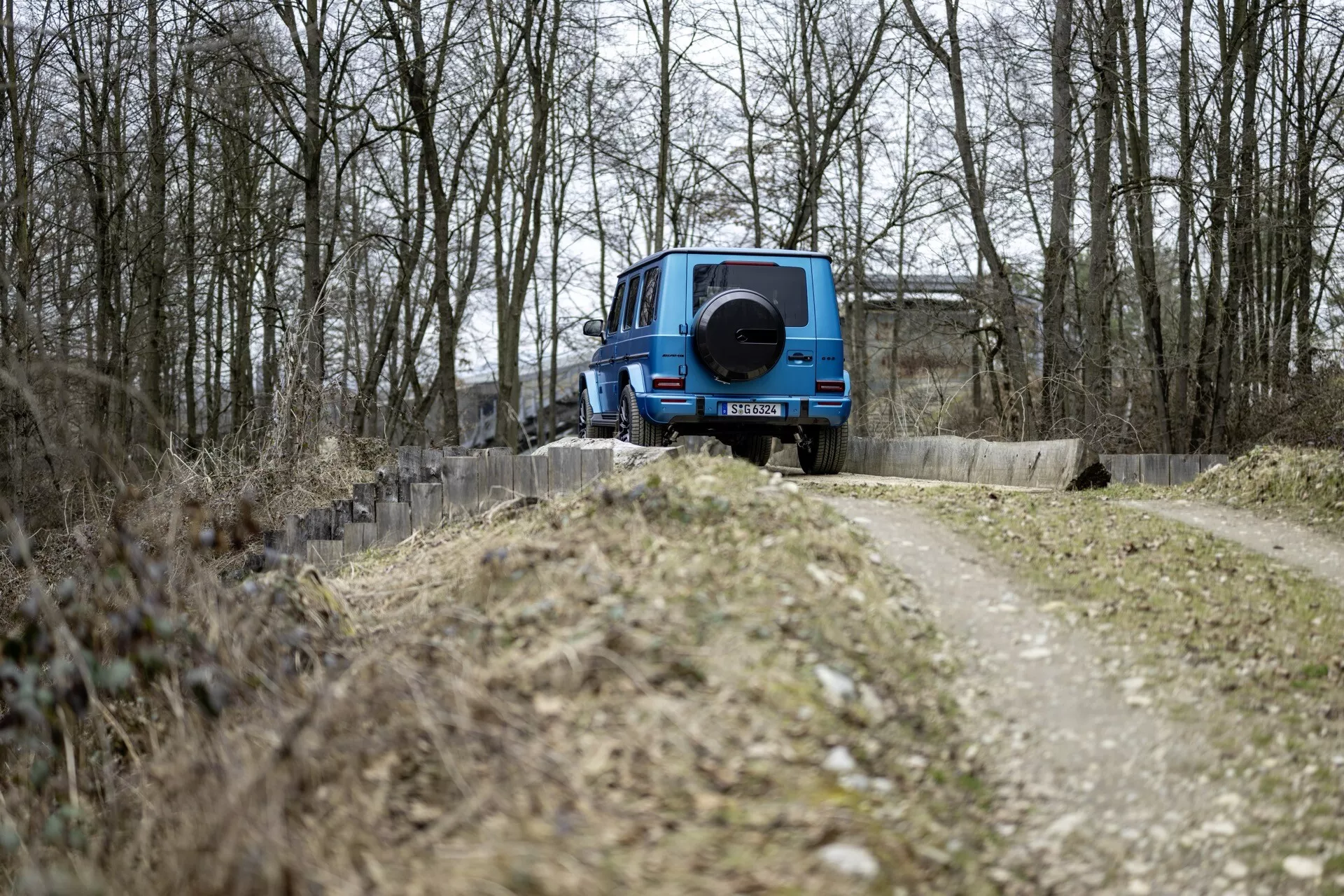 Niebieskie SUV Mercedes-Benz klasy G 2024, zaparkowane tyłem do kamery obok wąskiej drogi gruntowej otoczonej przez drzewa bez liści, prezentujące design off-road z kołem zapasowym zamontowanym na tyle, pod zachmurzonym światłem bez widocznych funkcji technologicznych na zewnętrznym tylnym.