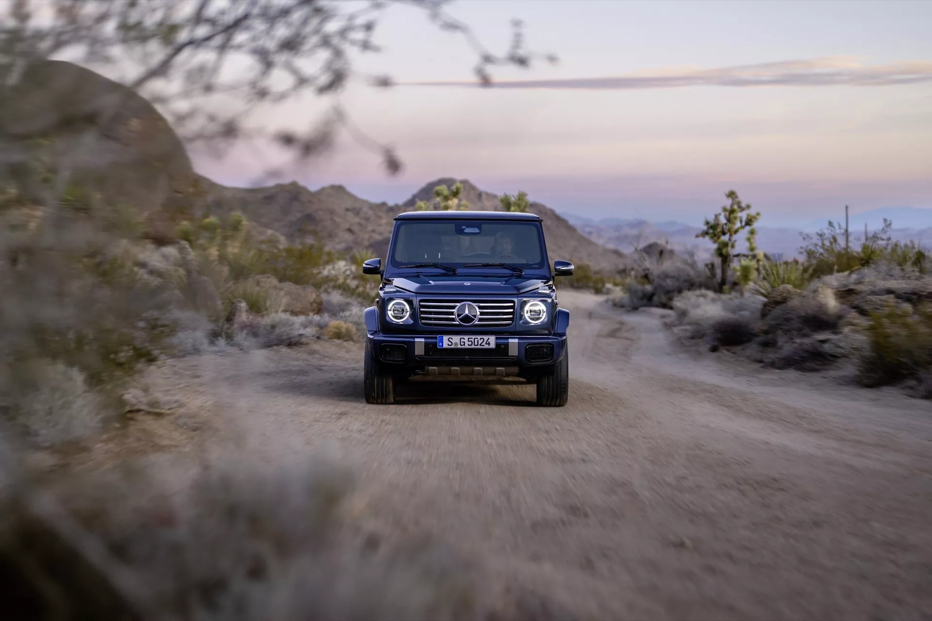 SUV Mercedes-Benz klasy G z 2024 roku jadący po drodze gruntowej z włączonymi światłami, podkreślający jego solidną konstrukcję i zdolności terenowe z widoku frontalnego o zmierzchu