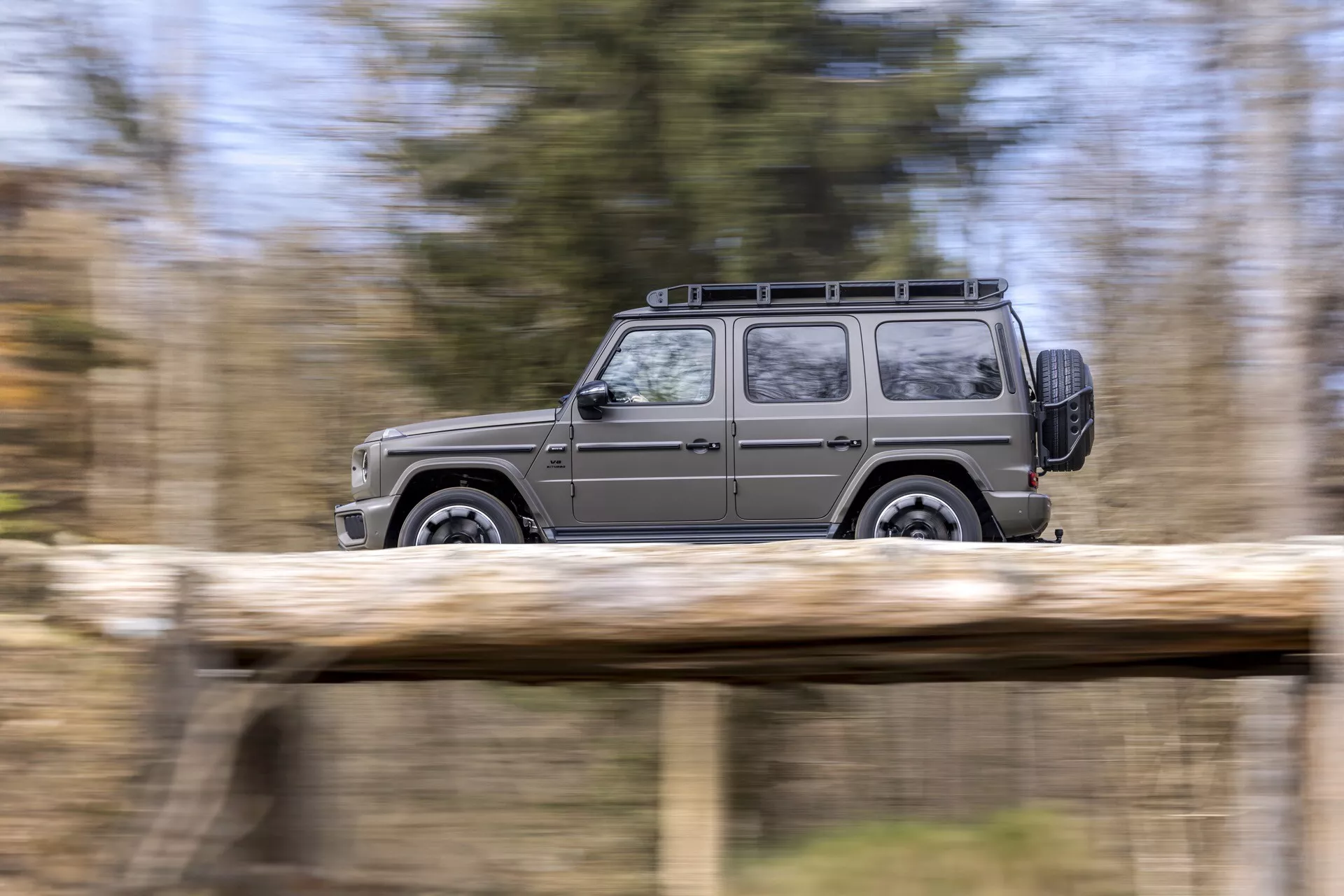 Dynamiczny profil boczny SUV Mercedes-Benz Klasy G (G-Klasse) 2024 ukazujący jego ruch z rozmazanym tłem naturalnym, podkreślający jego kanciasty design, bagażnik dachowy i koła ze stopów lekkich