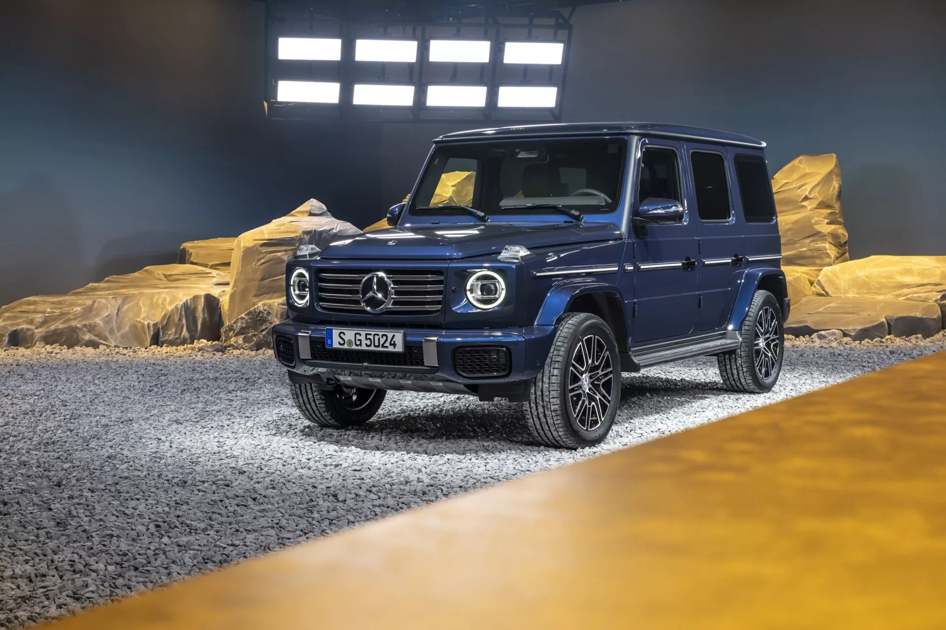 Luksusowy SUV Mercedes-Benz Klasa G (G-Klasse) rocznika 2024 w salonie z dramatycznym oświetleniem, przedstawiający ukośny widok z przodu i boku podkreślający okrągłe światła LED, dominującą gril z dużym emblematem, solidny przedni zderzak z zaawansowaną technologią, felgi z lekkich stopów z wieloma szprychami, oraz solidny, ale wyrafinowany design odpowiedni do użytku terenowego i miejskiego, z tablicą rejestracyjną 'S-GO524'.