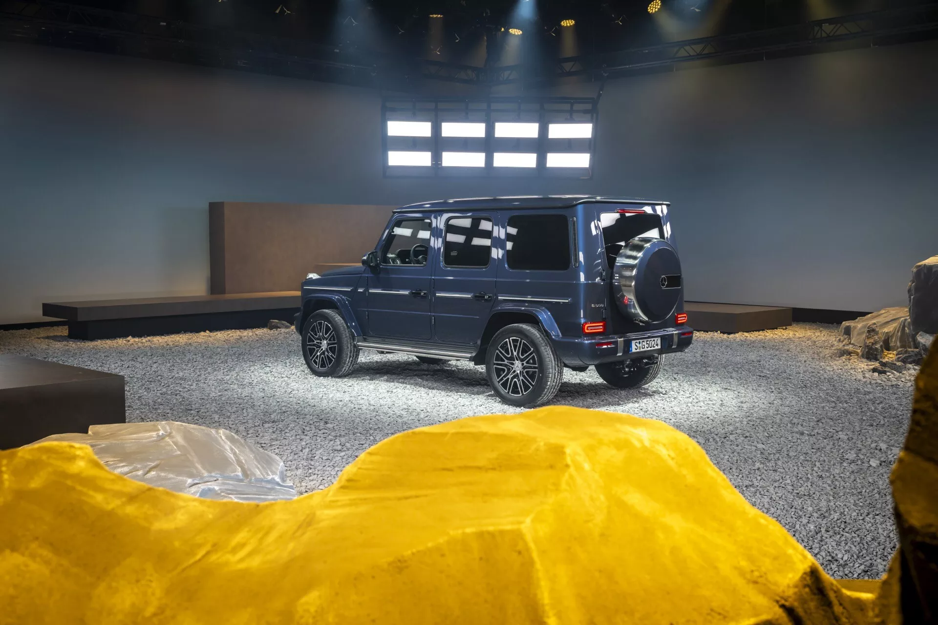 SUV Mercedes-Benz Klasa G (G-Klasse) 2024 w salonie, prezentujący widok z tyłu i boku z podkreślonymi zdolnościami terenowymi dzięki kołu zapasowemu zamontowanemu na tyle, kołom ze stopów i wysokiemu prześwitowi na zaaranżowanym tle