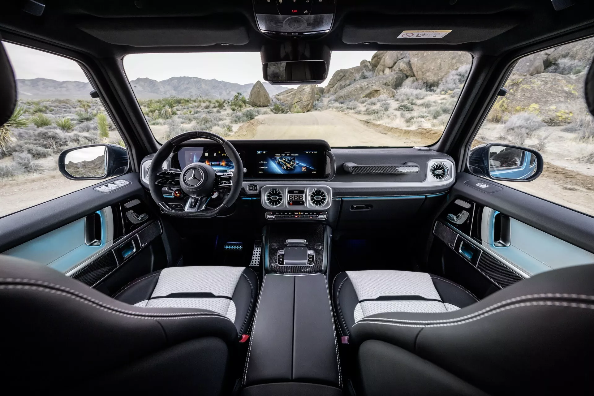 Widok wnętrza Mercedes-Benz Klasy G 2024 z tylnej kanapy, pokazujący deskę rozdzielczą, cyfrowy zestaw wskaźników, kierownicę owiniętą skórą z kontrolami, duży system infotainmentu z ekranem dotykowym, klimatyzację dwustrefową, wytrzymałą dźwignię zmiany biegów, przednie fotele kubełkowe z materiałów premium i panoramiczny dach.