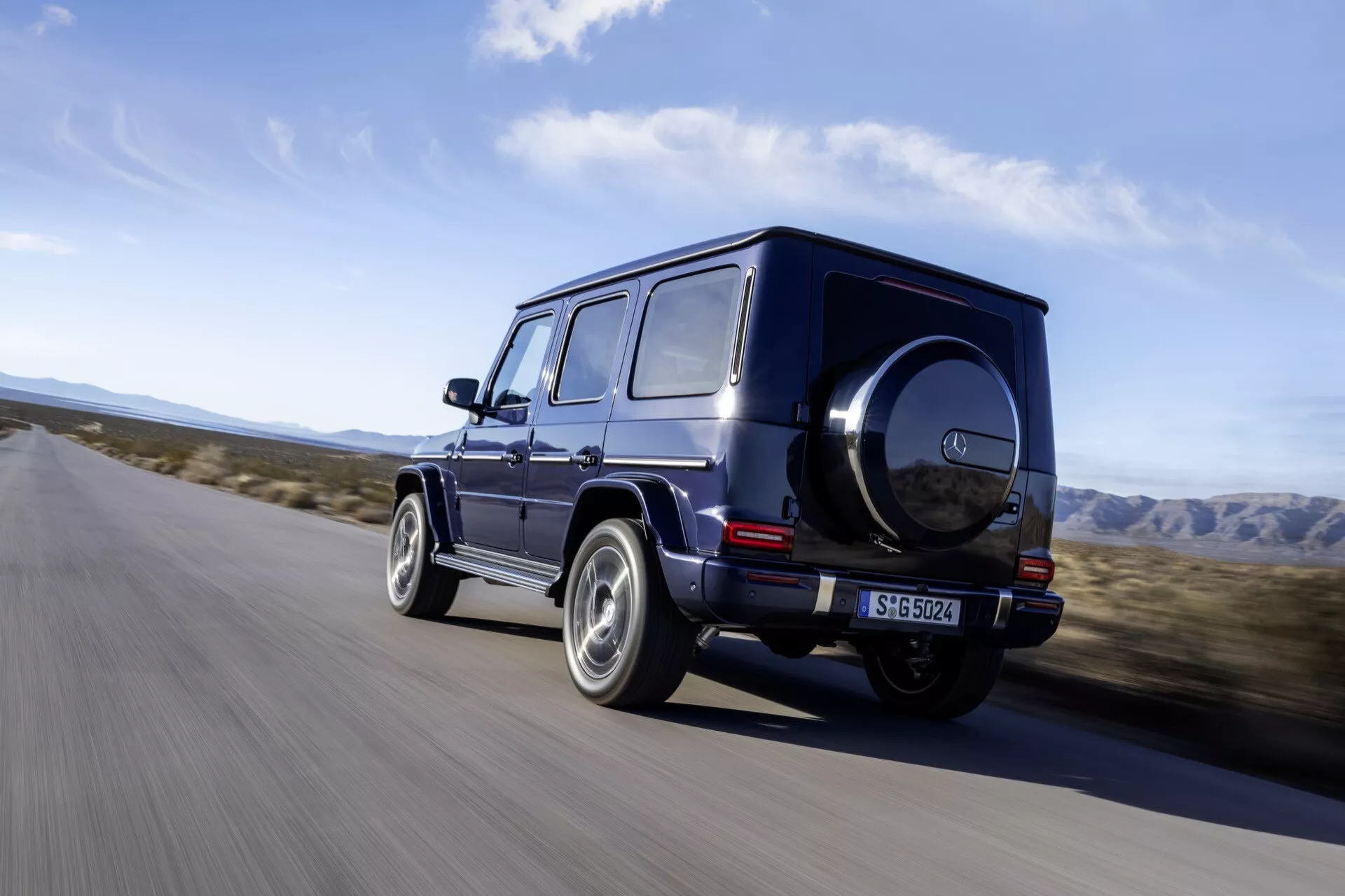 Luksusowy SUV Mercedes-Benz G-Klasse 2024 w ciemnym kolorze jadący po asfaltowej drodze w środowisku przypominającym pustynię, prezentujący widok z tylnej części z błyszczącą obręczą, kanciastym wzornictwem i cechami terenowymi, takimi jak koło zapasowe i tylne światła LED.