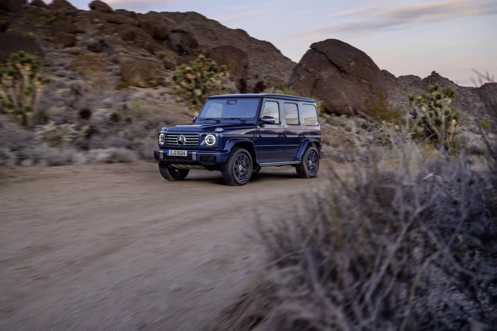 SUV Mercedes-Benz Klasy G 2024 jadący po nieutwardzonej drodze pustynnej, z reflektorami LED, technologią adaptacyjną, luksusowym wnętrzem, terenowym designem i zaawansowanymi funkcjami bezpieczeństwa, z rozmyciem ruchu podkreślającym dynamiczne możliwości