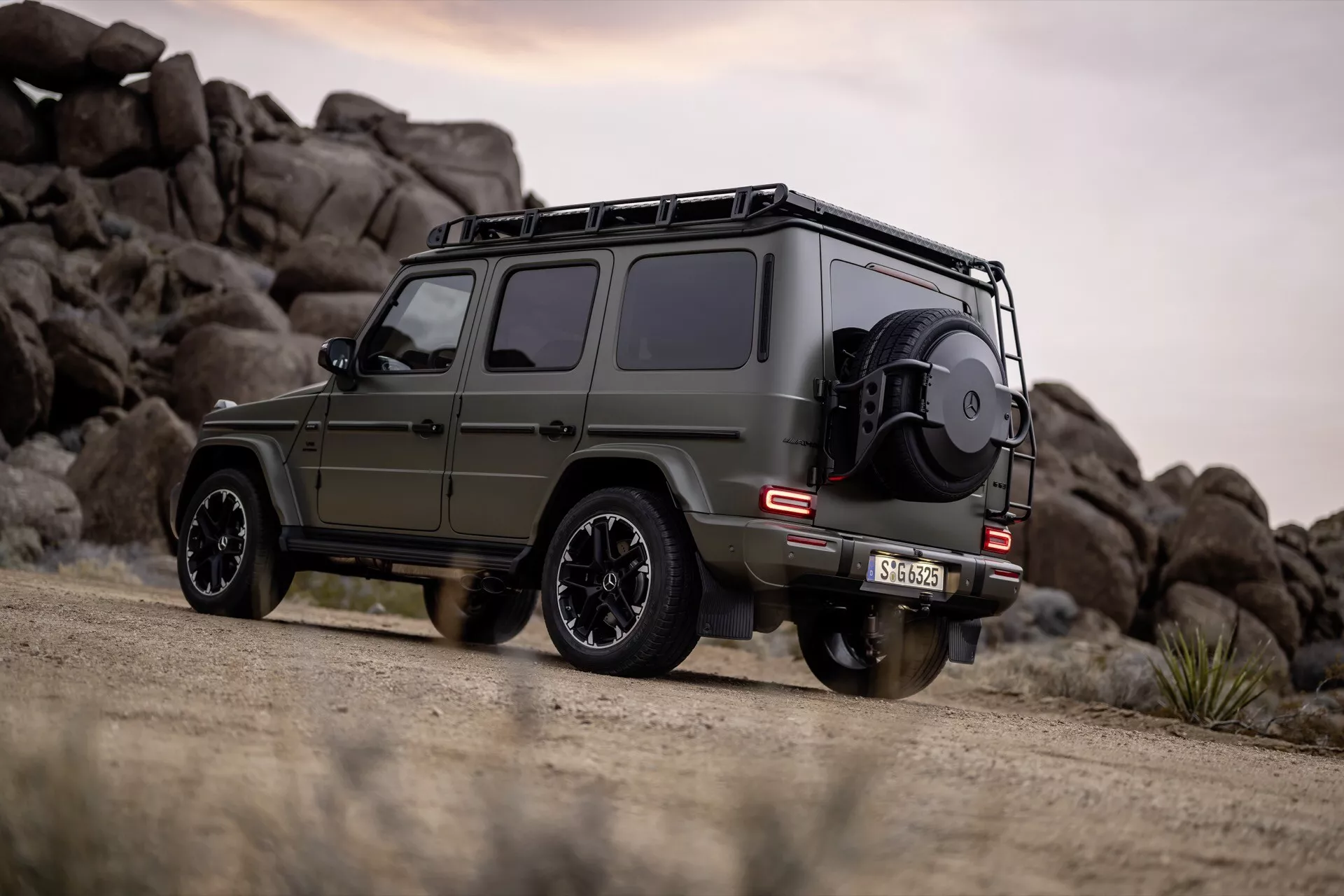 Mercedes-Benz Klasa G (G-Klasse 465) 2024 w matowym oliwkowo zielonym kolorze, przedstawiający widok boczny i tylny z funkcjami terenowymi, zaparkowany na drodze gruntowej na tle skalistego krajobrazu.