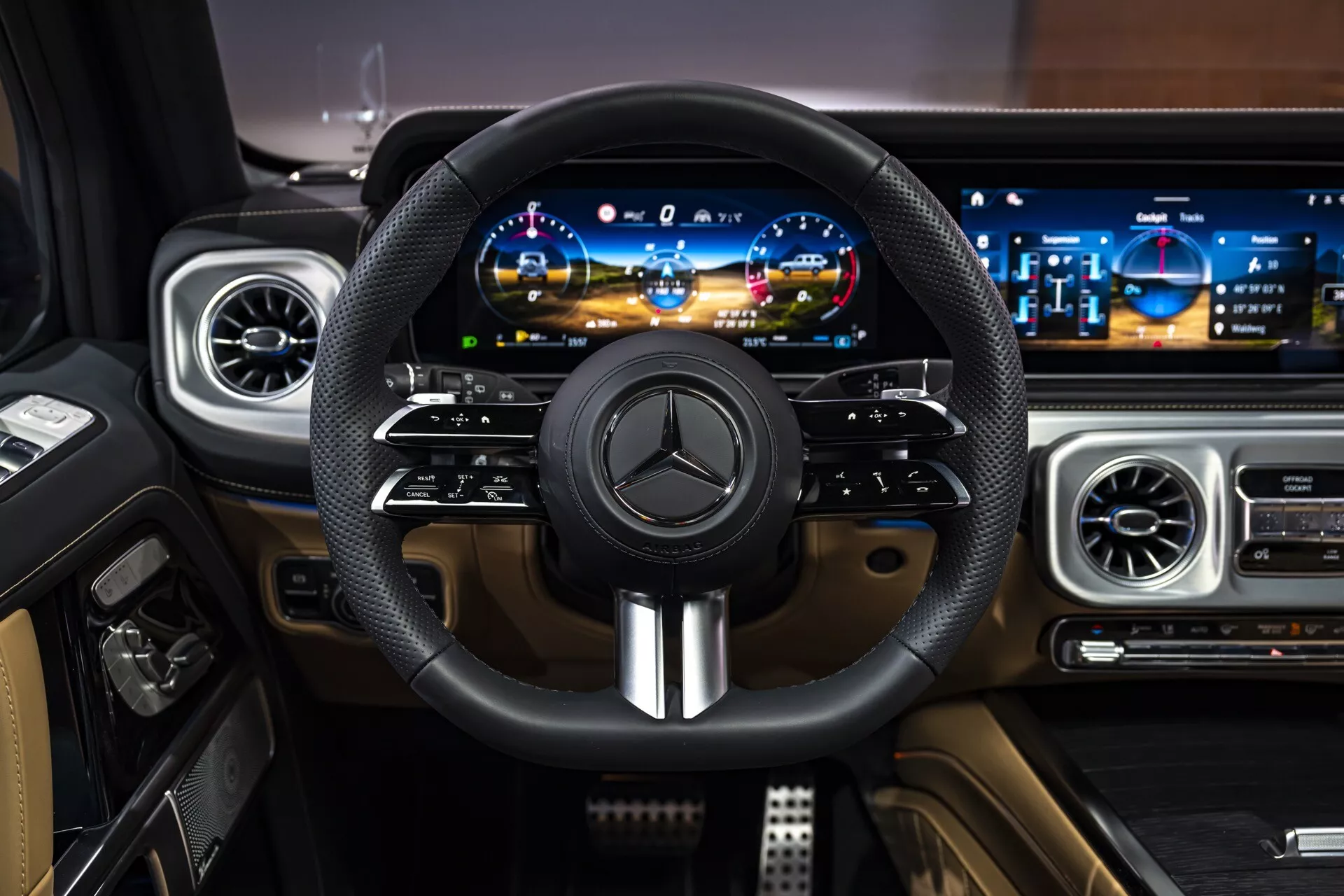 Wnętrze Mercedes-Benz Klasy G (G-Klasse) 2024 przedstawiające luksusowy kokpit z perspektywy kierowcy, zawierające kierownicę owiniętą skórą z emblematem Mercedes i metalicznymi akcentami, cyfrowy panel przyrządów i szerokoekranowy panel infotainment