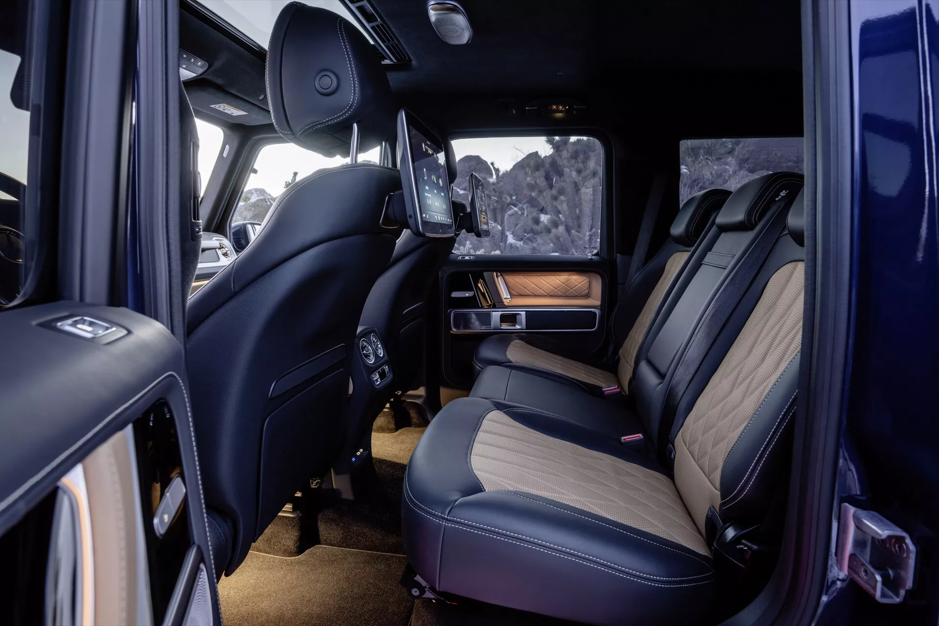 Widok wnętrza Mercedes-Benz Klasy G 465 2024 ukazujący tylną kanapę premium, szczegółowe szycie, luksusowe materiały z akcentami z drewna, zaawansowane funkcje technologiczne, w tym wysuwany ekran rozrywki, oświetlenie ambientowe i kontrolę klimatu z perspektywy otwartych drzwi pasażera