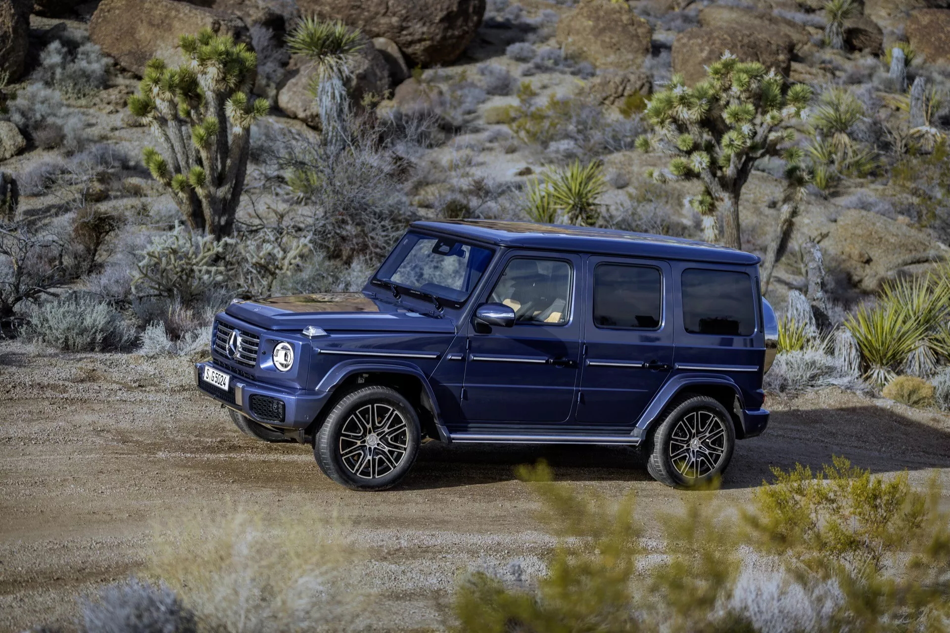 Ciemny SUV Mercedes-Benz Klasa G 465 z 2024 roku, zaparkowany w terenie na nieutwardzonej drodze z otoczeniem suchych roślinności, prezentujący jego solidną, kwadratową konstrukcję, duże, wieloszprychowe koła oraz relingi dachowe odpowiednie do przygód i luksusowych zdolności terenowych.