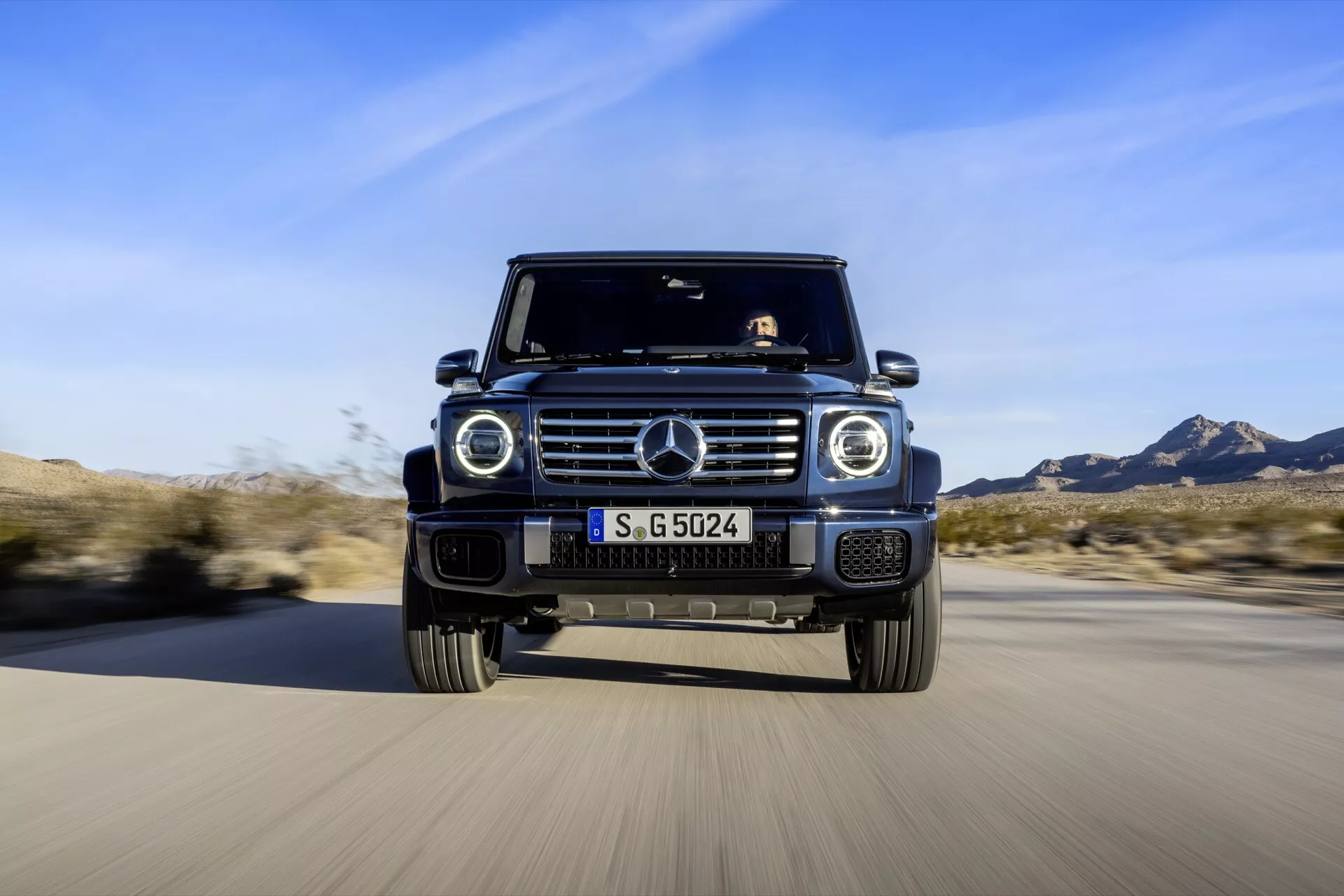 SUV Mercedes-Benz Klasy G 2024 zbliżający się na drodze, pokazujący niski kąt widzenia jego emblemowej kraty, okrągłych świateł LED, płyty osłaniającej na przednim zderzaku, przedniej kamery i jego charakterystycznej, kanciastej sylwetki na rozmytym tle, podkreślając dynamiczną obecność i zaawansowane funkcje pojazdu.