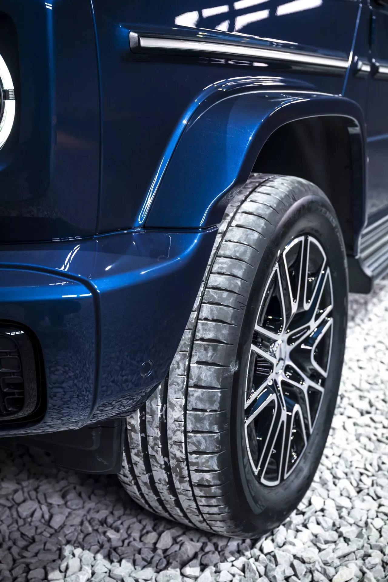 Zbliżenie na tylną część koła i karoserię Mercedes-Benz klasy G 2024 w kolorze niebieskim, pokazujące koło z felgą aluminiową o wielu szprychach, wyraźny bieżnik opony i charakterystyczne nadkola sugerujące wysoką jakość lub zdolności terenowe