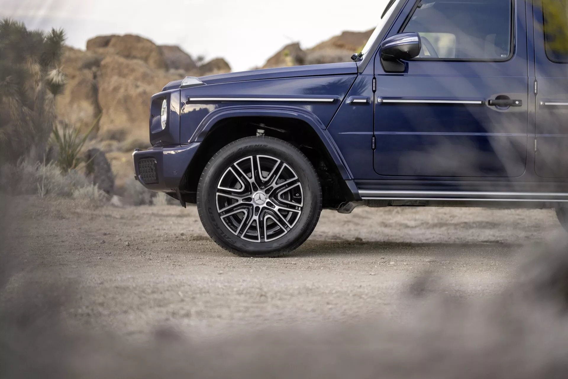 Zbliżenie na przednie koło i błotnik od strony kierowcy Mercedes-Benz G-Klasse 2024 w kolorze ciemnoniebieskim, ukazujące luksusowe koła ze stopu i profil boczny sugerujący zdolności terenowe