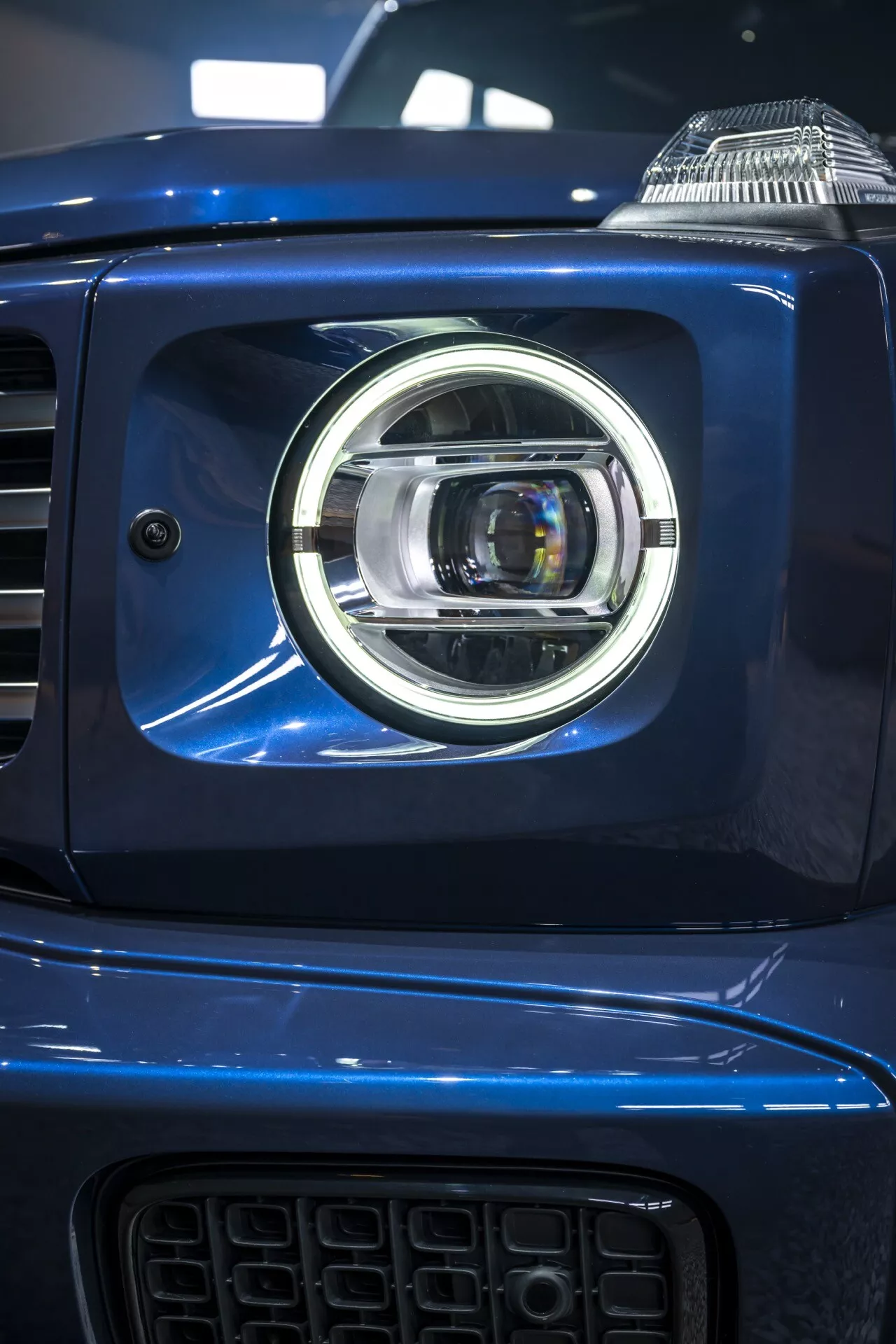 Zbliżenie na lewy zespół reflektorów Mercedes-Benz G-Klasse 2024, przedstawiający nowoczesny projekt reflektora LED z chromowanymi akcentami na metalicznie niebieskim nadwoziu