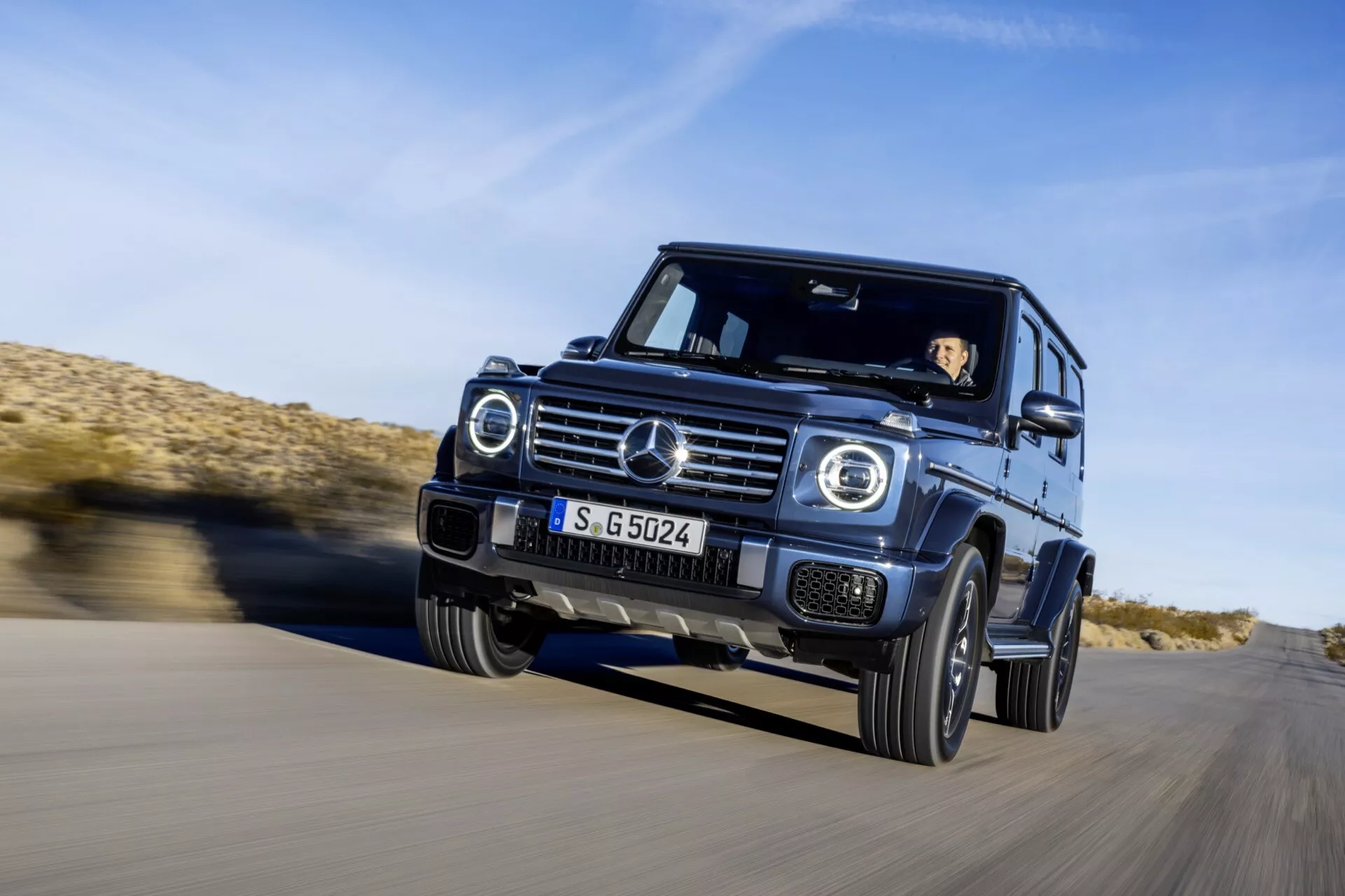 SUV Mercedes-Benz Klasy G 2024 jadący po asfalcie w środowisku pustynnym, widok z przodu pod kątem trzech czwartych ukazujący dynamiczny ruch, reflektory LED, chromowaną atrapę i zdolności terenowe