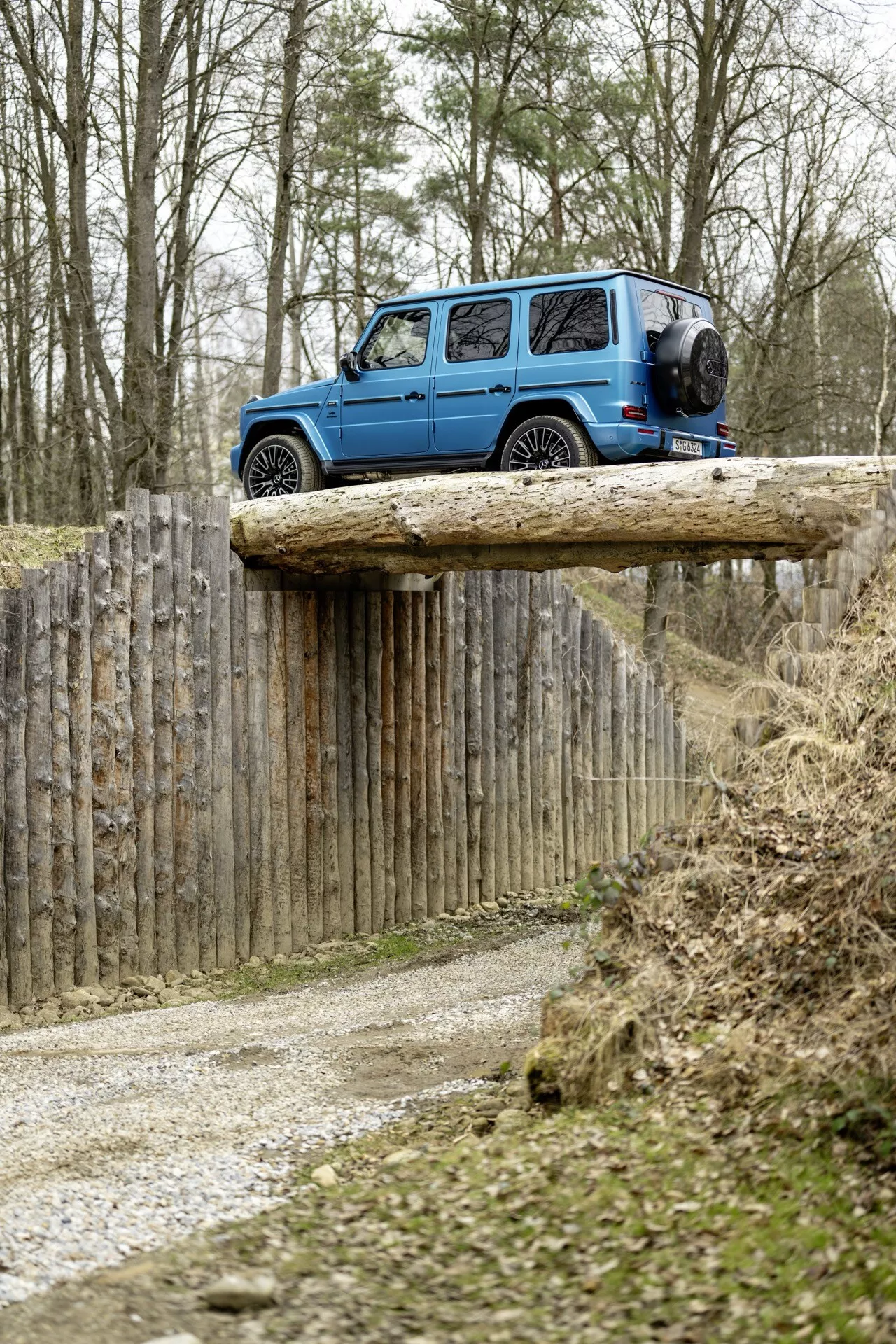 Niebieski SUV Mercedes-Benz G-Klasse 2024 na podwyższonej drewnianej platformie w terenie, pokazujący widoki z tyłu i boku z widocznym kołem zapasowym