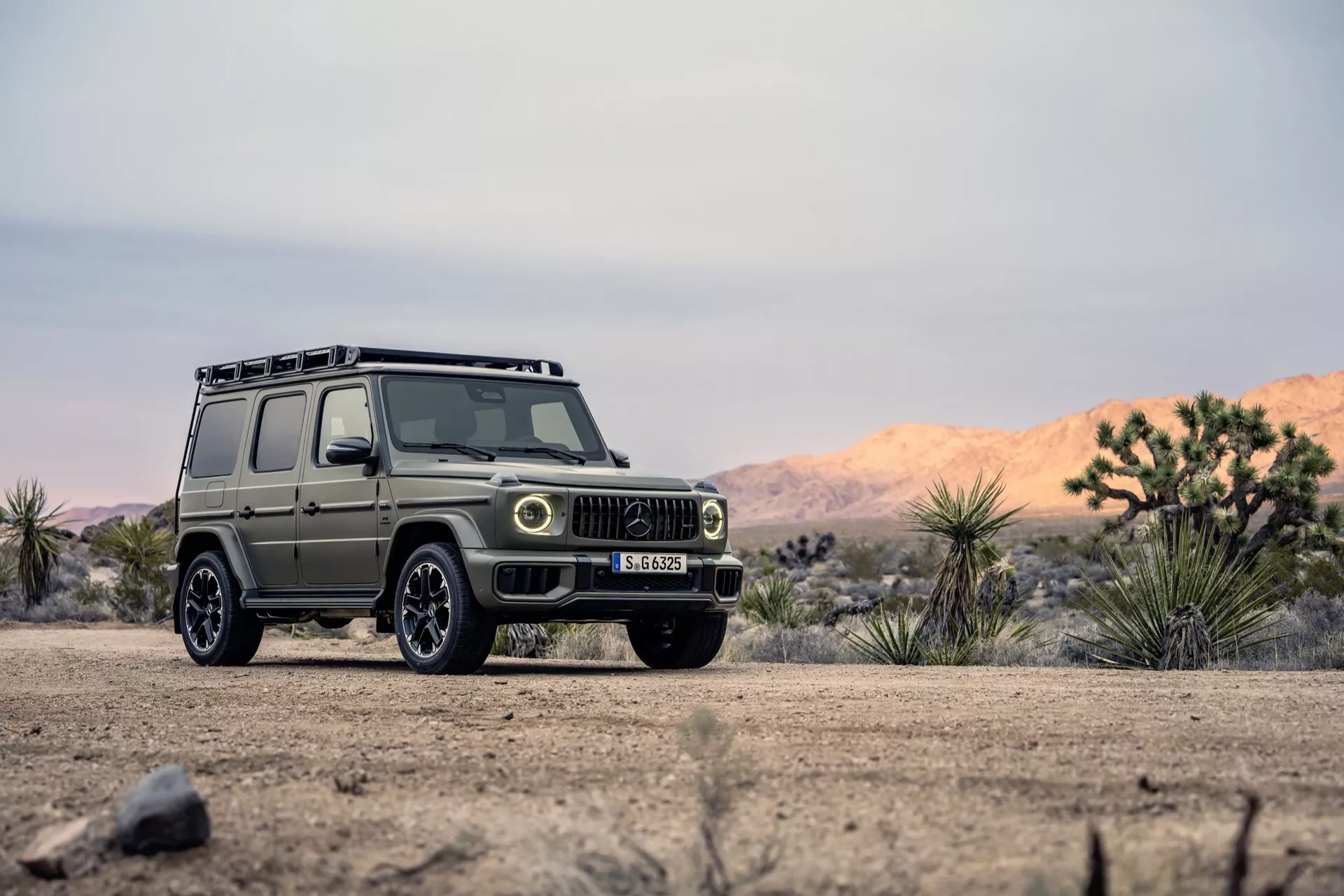 SUV Mercedes-Benz G-Klasse 2024 w surowym terenie zewnętrznym, pokazujący widok z przedniego trójkąta z detalami grilla, reflektorów i kół terenowych na drodze gruntowej