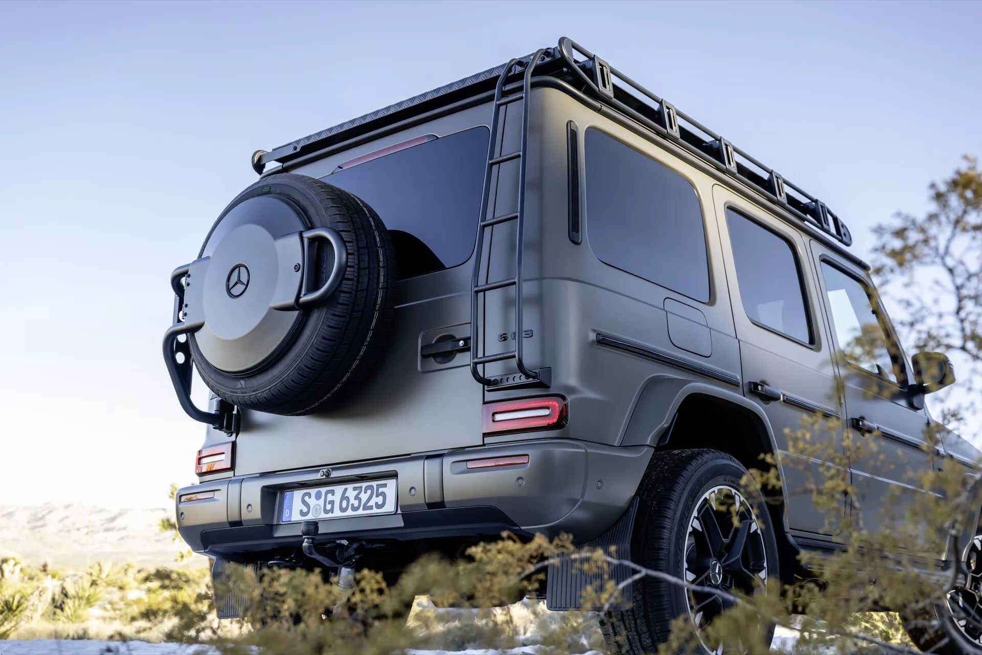Widok z tyłu pod kątem trzech czwartych SUV-a Mercedes-Benz G-Klasse 2024 pokazujący prawą stronę, zdjęcie z niskiego kąta podkreślające potężną posturę z zaakcentowanymi detalami, w tym wąskie pionowe światła tylne LED, koło zapasowe zamontowane na tylnych drzwiach z pokrowcem z emblematem, solidną, pudełkową konstrukcję wskazującą na zdolności terenowe, czujniki parkowania, hak holowniczy oraz bagażnik dachowy na metalicznym wykończeniu pod oświetleniem ambientalnym.