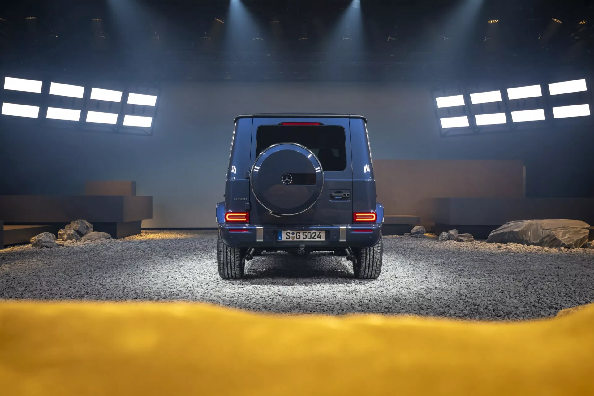 Tylny widok SUV-a Mercedes-Benz Klasy G (G-Klasse) 2024 w studiu imitującym teren off-roadowy pokazujący wysokość, tylne światła, koło zapasowe i strategiczne oświetlenie
