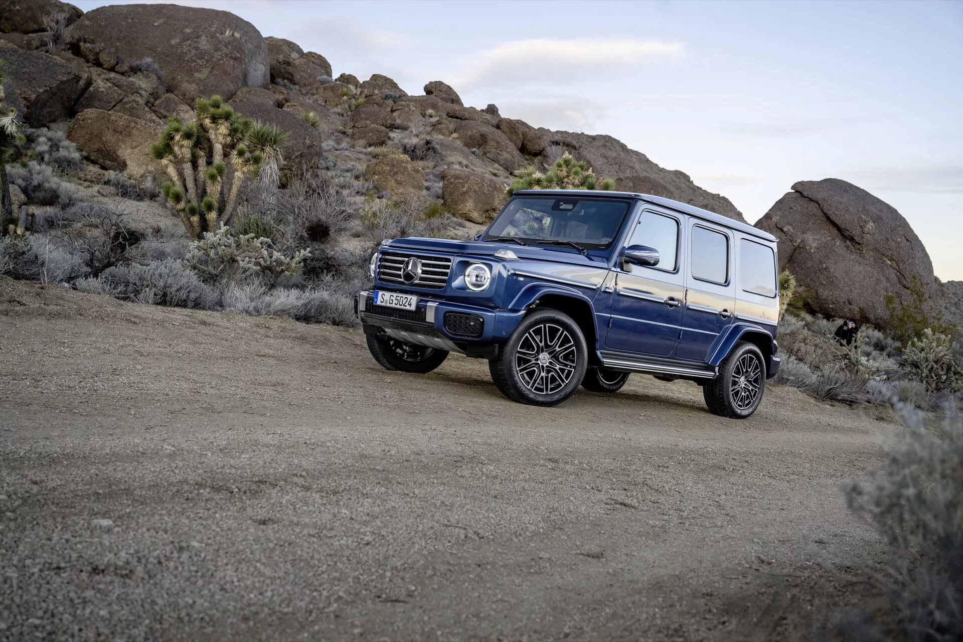 Niebieskie SUV Mercedes-Benz Klasy G 465 z 2024 roku zaparkowane na nierównym terenie, prezentujące kwadratową konstrukcję, dużą prześwit i zdolności terenowe pojazdu z perspektywy trzech czwartych przedniej, podkreślające alufelgi i opony terenowe