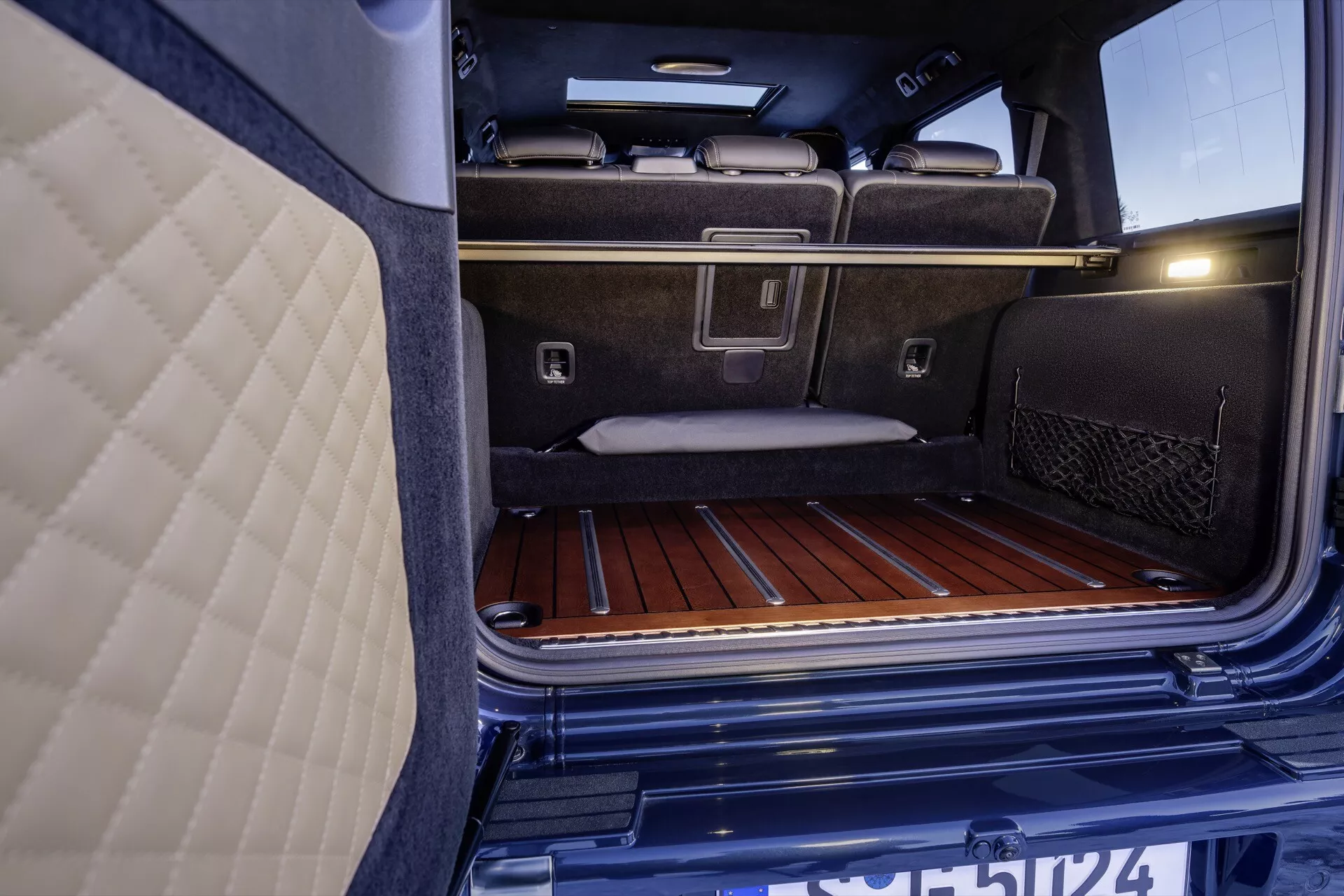 Zbliżony widok wnętrza bagażnika Mercedes-Benz G-Klasse 2024, podkreślający jego luksusową wykładzinę dywanową, listwy podłogowe z drewna, metalowe kotwice do mocowania ładunku i kieszeń siatkową, obok luksusowych tylnych siedzeń i zintegrowanego oświetlenia bagażnika