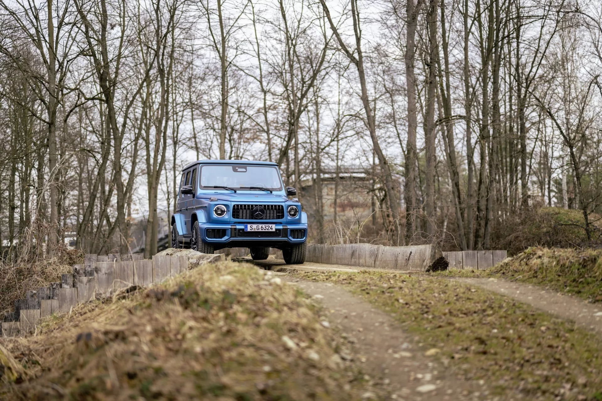 Niebieski pojazd terenowy Mercedes-Benz G-Klasse 2024 w ruchu na drodze gruntowej, widok z przodu pod kątem trzech czwartych pokazujący grill przedni, maskę, przednią szybę i wysokie prześwit, w leśnym otoczeniu bez śniegu