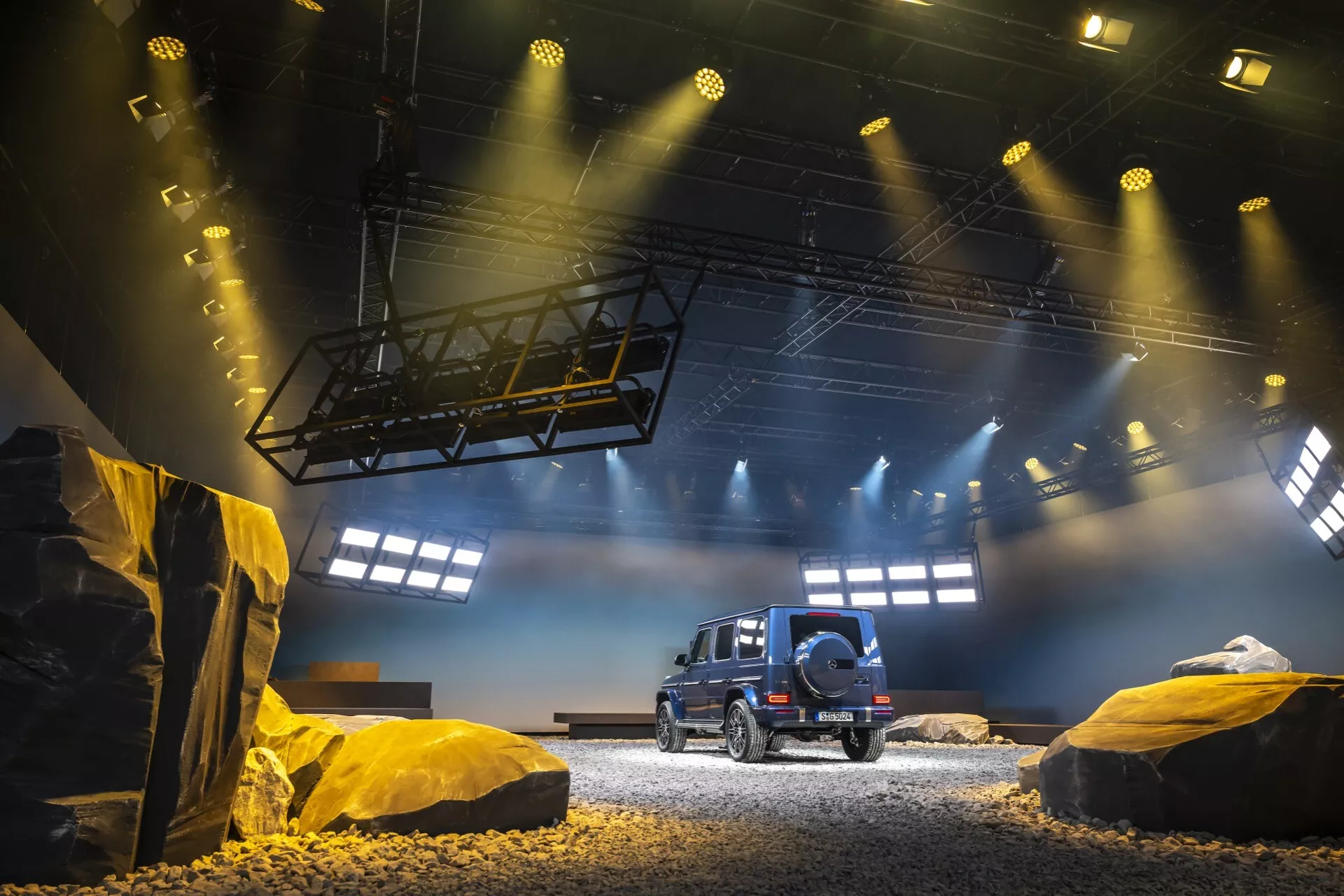 SUV Mercedes-Benz G-Klasse 2024 wystawiony w symulowanym terenie off-road, prezentujący widok pojazdu z tyłu i boku z detalami profilu bocznego, tylnej szyby, tylnych świateł LED oraz zapasowego koła zamontowanego na tylnej klapie, pod dramatycznym oświetleniem wystawienniczym