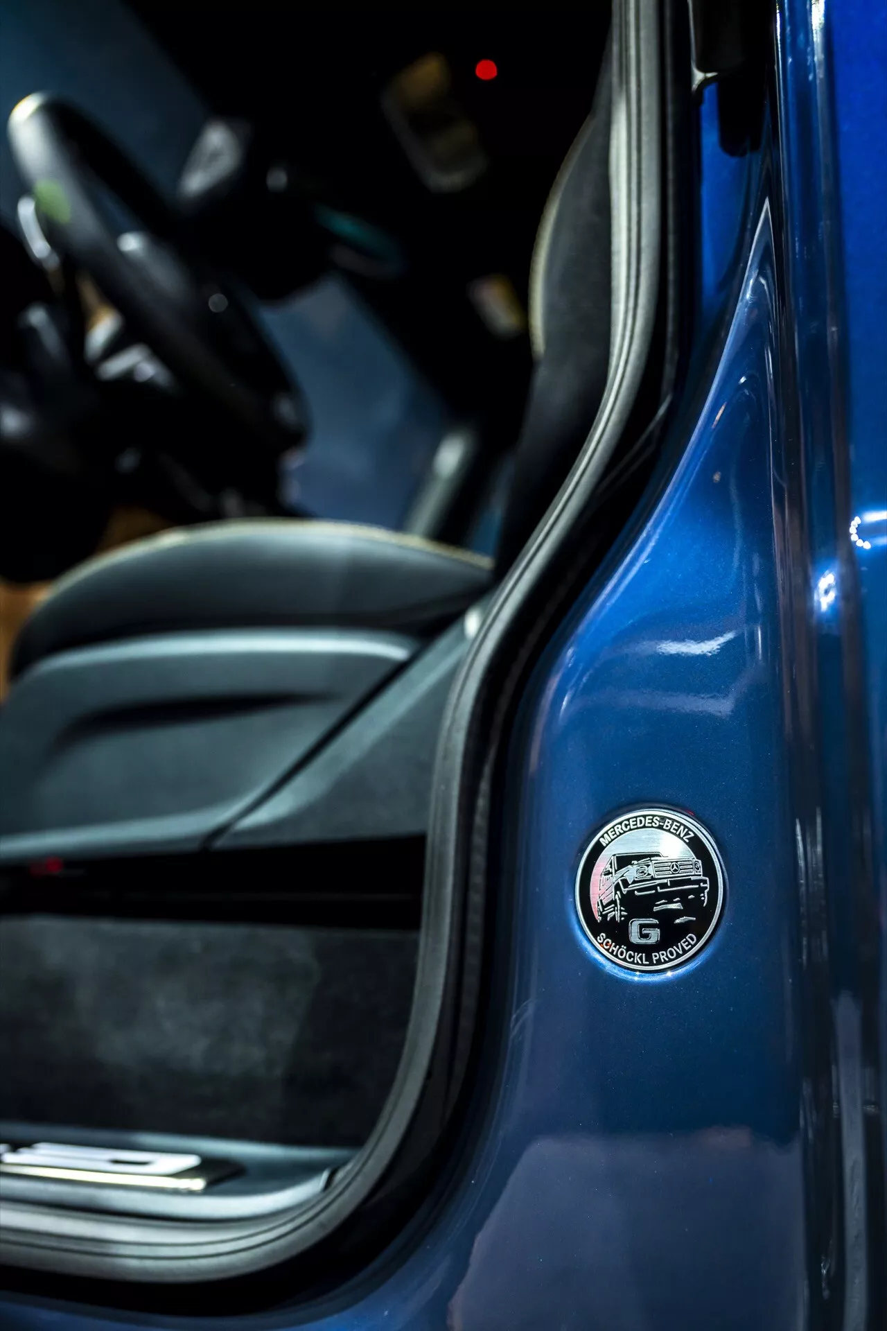 Zbliżenie przedstawiające detal emblematu na słupku B Mercedesa-Benz G-Klasse 2024, z jaskrawo niebieskim nadwoziem i sportowym czarnym wnętrzem, podkreślające cechy pojazdu premium ze szczególnym uwzględnieniem marki i materiałów wysokiej jakości.