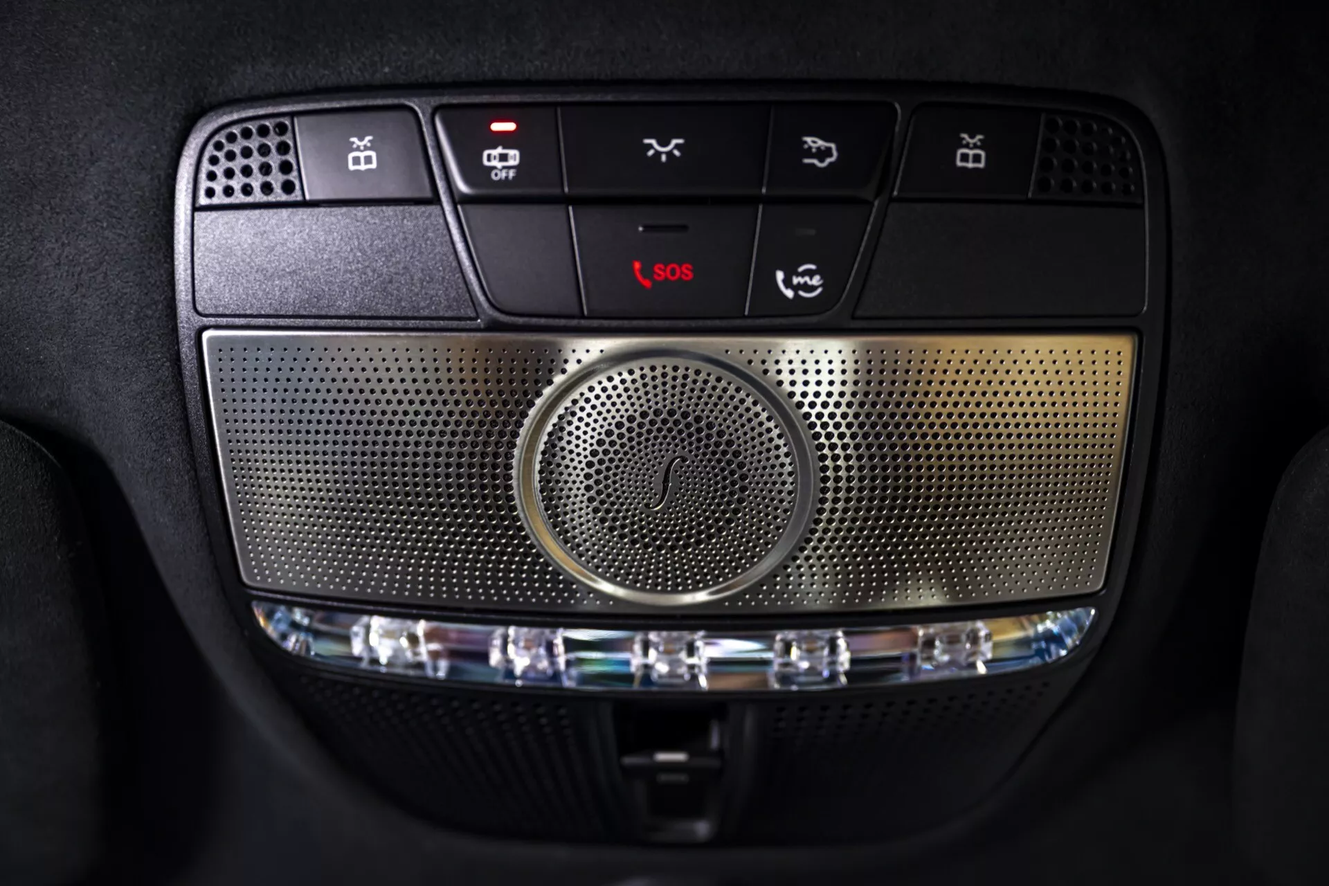Zbliżone zdjęcie konsoli sufitowej Mercedes-Benz G-Klasse 2024 (465) z przyciskami sterującymi, centralnym głośnikiem, opcjami oświetlenia wnętrza, usługami ratunkowymi i funkcjami dostosowanymi do użytkownika, podkreślające zaawansowany system multimedialny i eleganckie detale konstrukcyjne