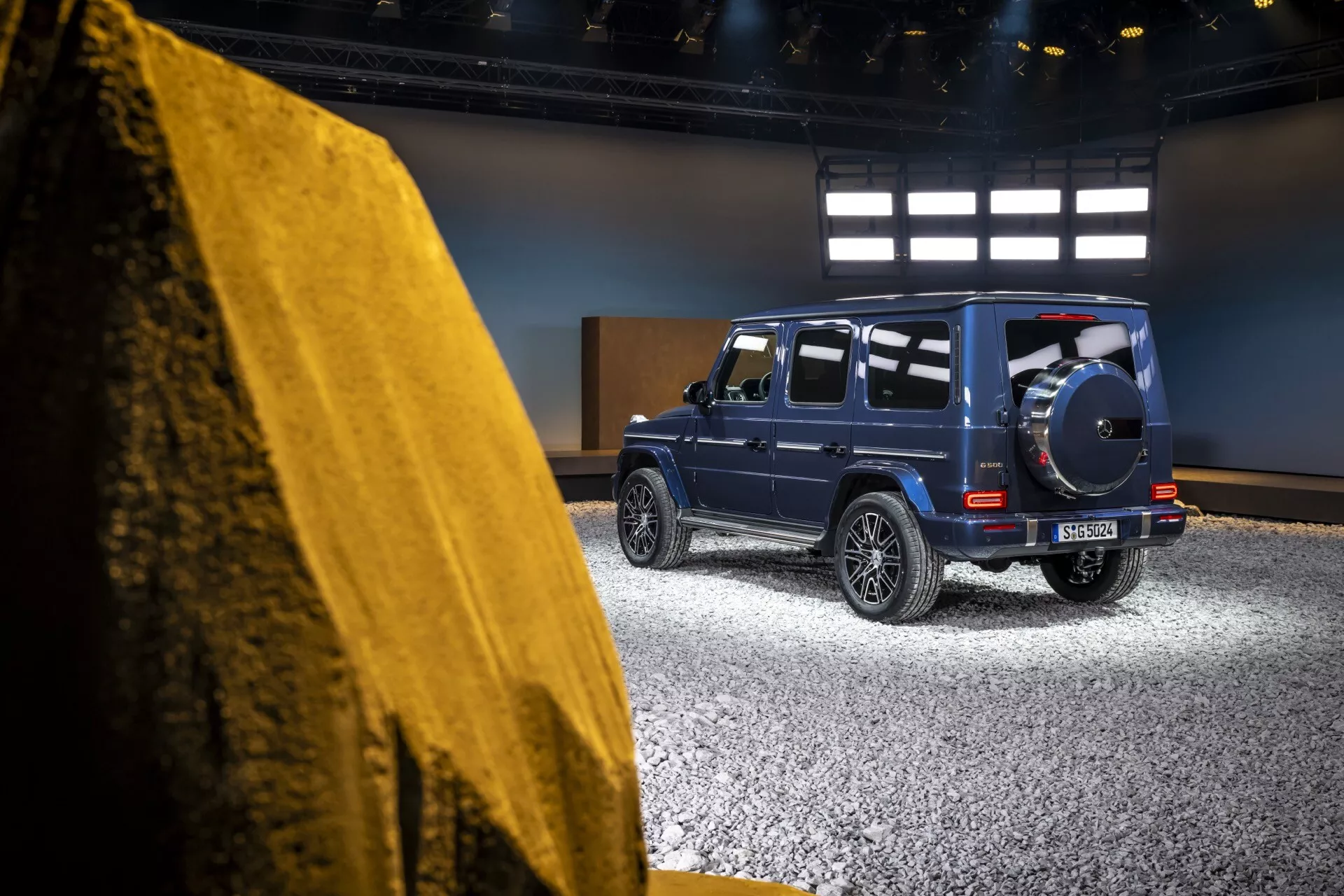 Boczny tylny widok Mercedes-Benz Klasy G (G 465) z 2024 roku w ustawieniu studyjnym, pokazujący jego sztywny luksusowy design, tylne światła LED oraz koło zapasowe na głębokim metalicznym niebieskim
