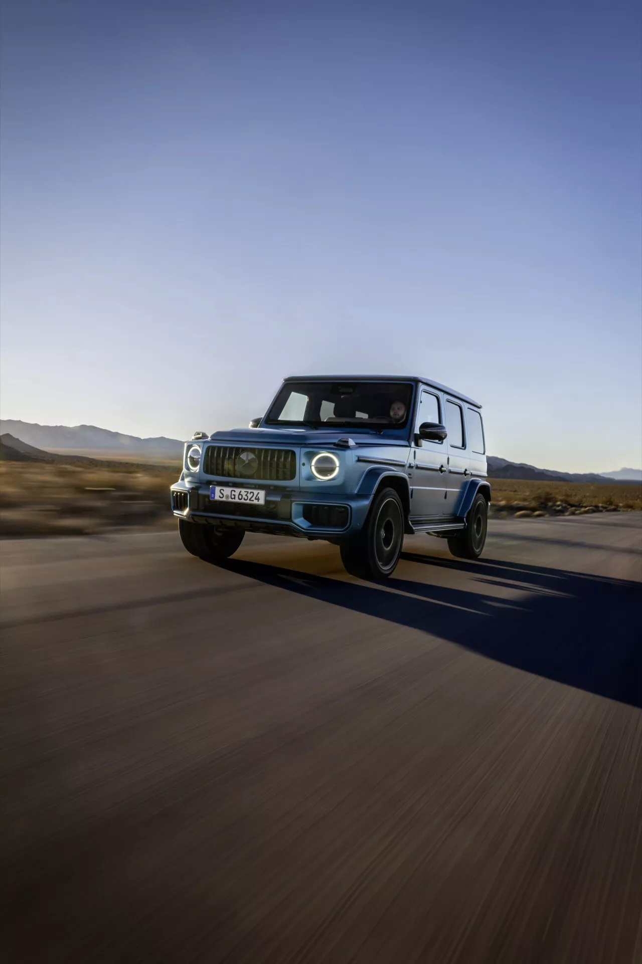 Mercedes-Benz G-Klasse 2024 w ruchu, prezentujące jego luksusowy design i funkcje z niskiego przedniego i bocznego kąta, podkreślające reflektory LED, grill przedni, felgi aluminiowe, sugerujące jego możliwości jazdy z wysoką prędkością i potencjał terenowy na pustynnym tle.