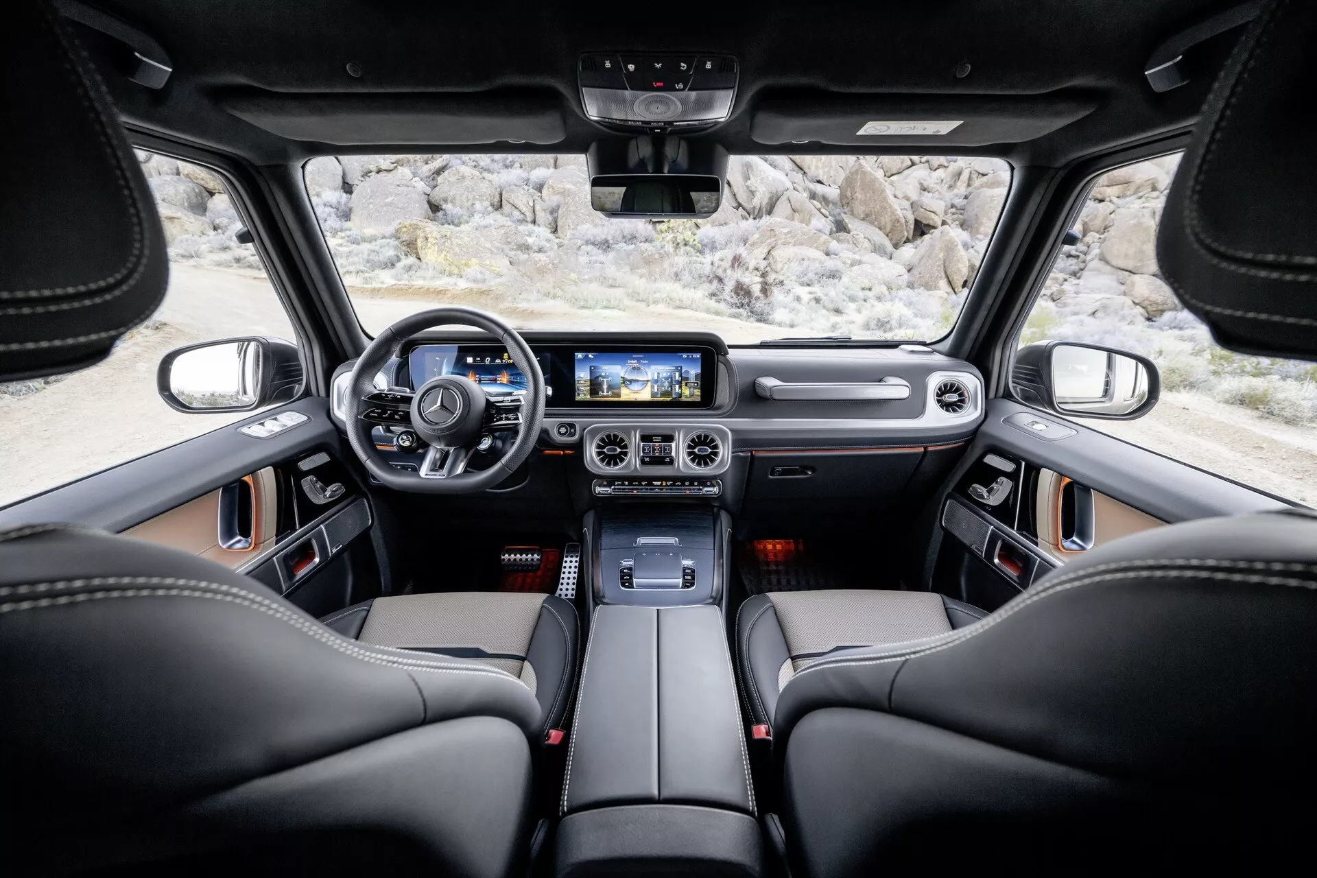 Wnętrze Mercedes-Benz Klasy G (G-Klasse 465) z 2024 roku z perspektywy tylnej kanapy pokazujące czarne skórzane fotele, nowoczesną kierownicę z przyciskami, deske rozdzielczą z elementami skórzano-metalowymi, cyfrowy kokpit, konsolę środkową z metalicznymi akcentami, duży ekran dotykowy systemu infotainment, pokrętła do kontroli klimatyzacji z metalicznym wykończeniem, kontrolki oświetlenia nad głową, zaawansowany system głośnikowy, panoramiczne okno dachowe, centralnie umieszczone lusterko wsteczne oraz przestronne okna zwiększające widoczność i efekt luksusu.