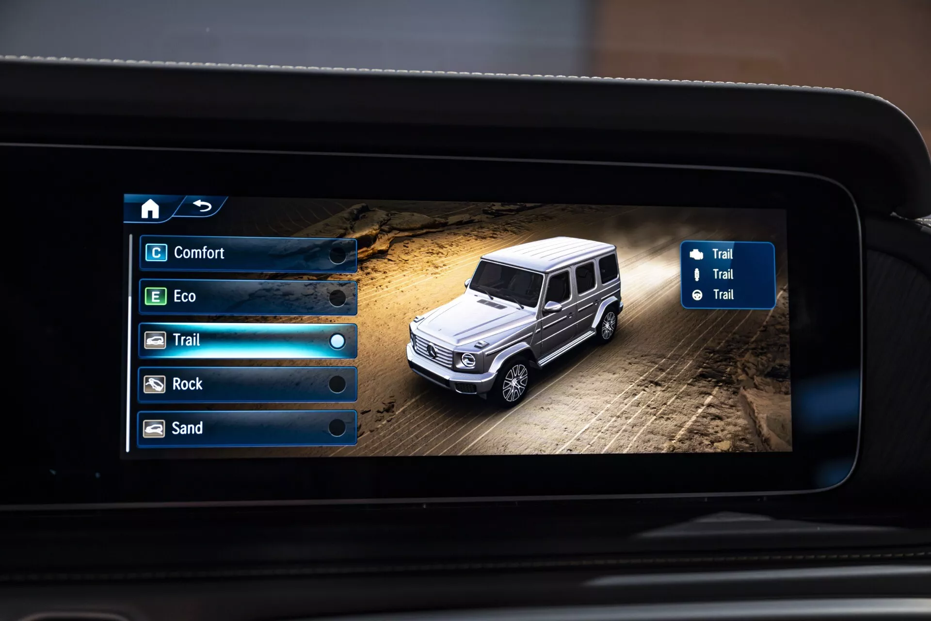 Cyfrowy wyświetlacz Mercedes-Benz klasy G (G-Klasse) z 2024 roku w terenowych warunkach pokazujący wybieralne tryby jazdy na ekranie systemu informacyjno-rozrywkowego, widok z lotu ptaka na luksusowego SUV-a na zakurzonym terenie z nawigacją i opcjami trybu