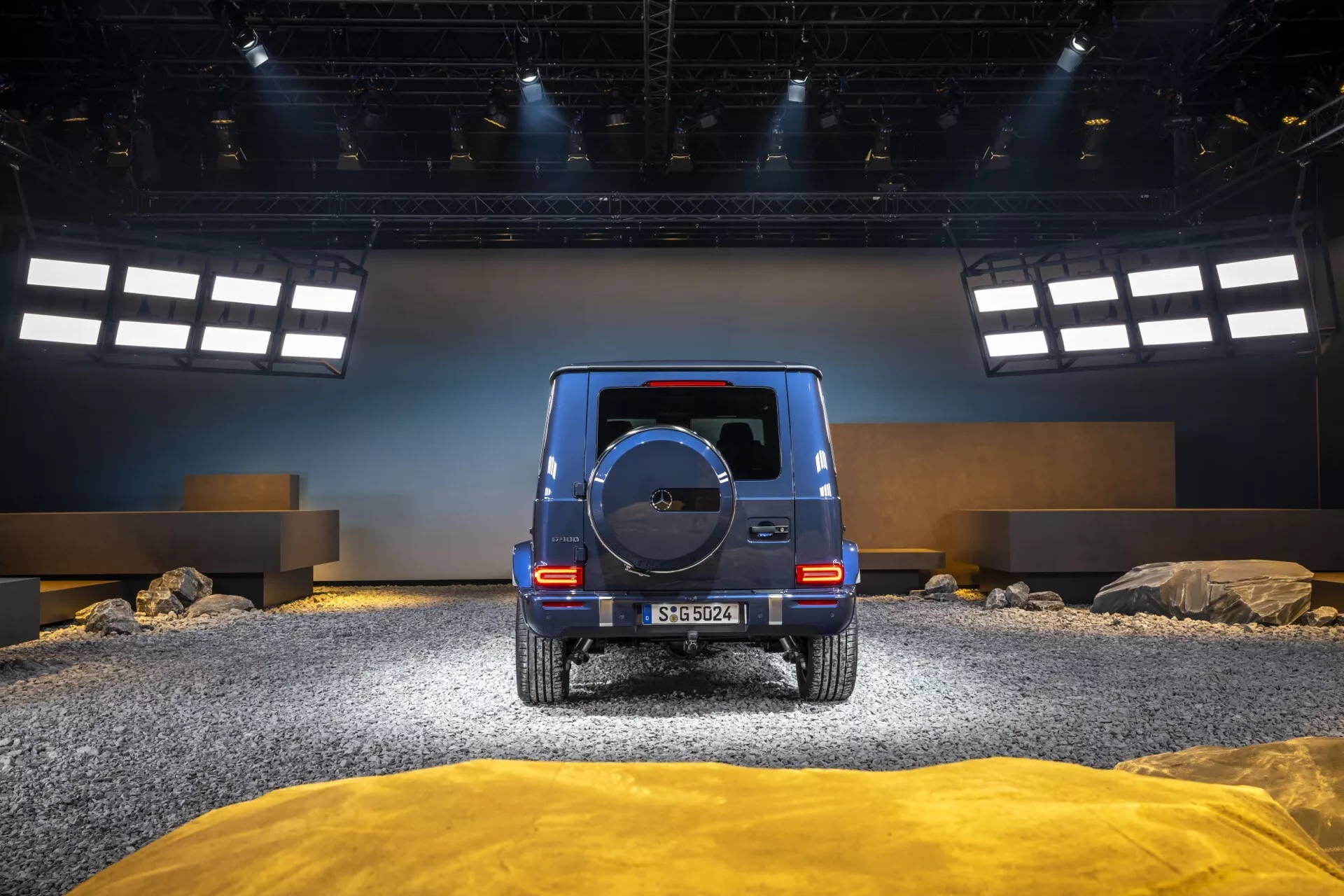 Tylny widok Mercedes-Benz Klasy G (G 465) 2024 w zaaranżowanym terenie off-road z kamieniami i teksturowanym podłożem, prezentuje tylne światła LED, koło zapasowe i błyszczące wykończenie.