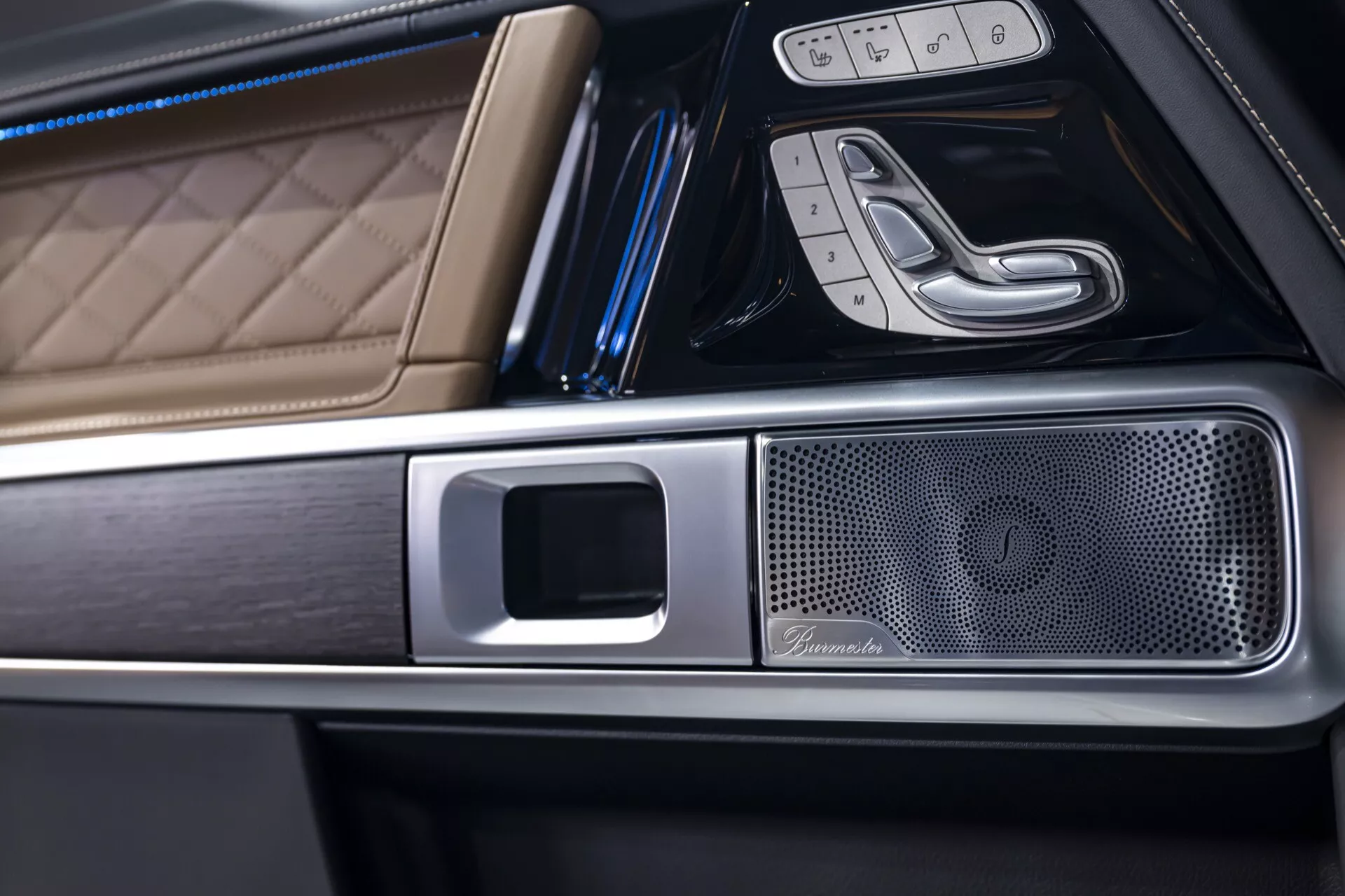 Zbliżenie wnętrza Mercedes-Benz G-Klasse 2024, podkreślające luksusową skórę z przeszyciami w kształcie diamentu, metaliczne wykończenia oraz wysokiej klasy kratkę głośnika na konsoli środkowej pod skośnym kątem