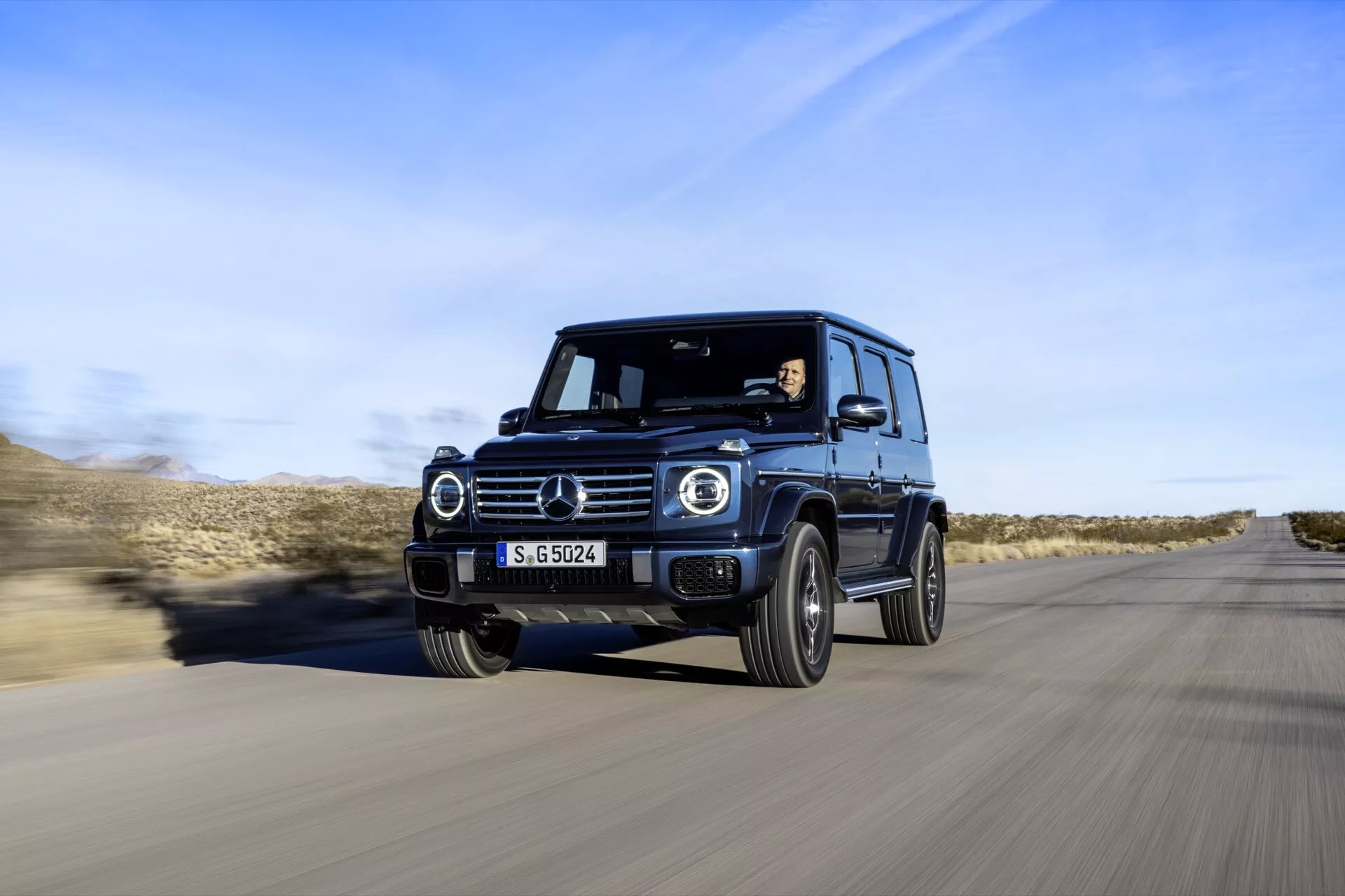 Luksusowy SUV Mercedes-Benz Klasy G 2024 w ruchu, uchwycony z przedniego kąta trzy czwarte, prezentujący reflektory LED, okrągłe światła do jazdy dziennej, solidny design i zaawansowane funkcje technologiczne na tle pustynnej drogi