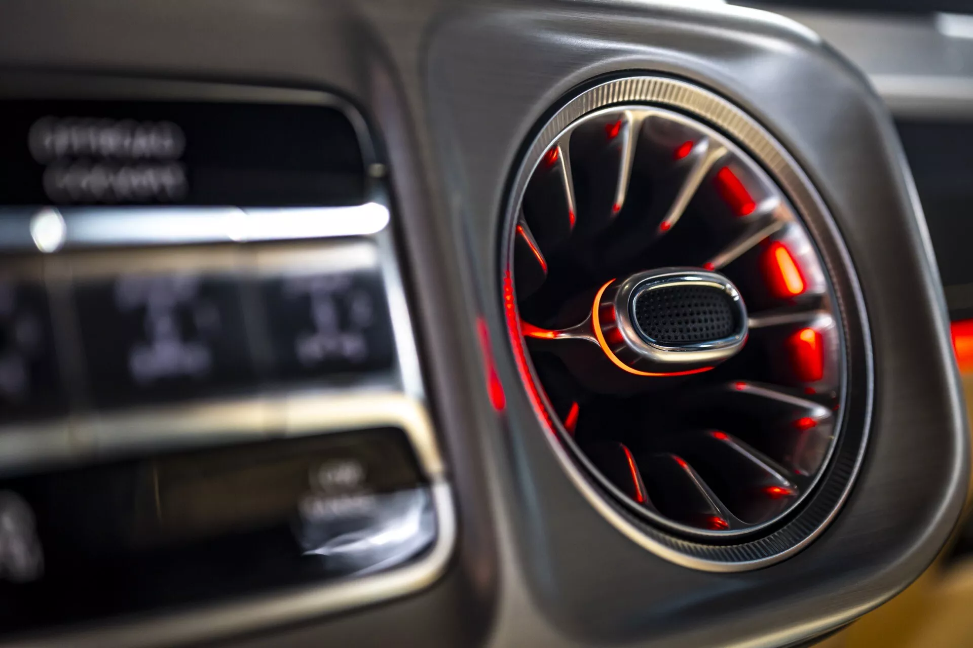 Zbliżenie na wnętrze Mercedes-Benz G-Klasse 2024 (465) ukazujące wywietrznik z wykończeniem chromowym i czerwonym oświetleniem ambientalnym oraz rozmytym tłem cyfrowego wyświetlacza podkreślające luksus i design
